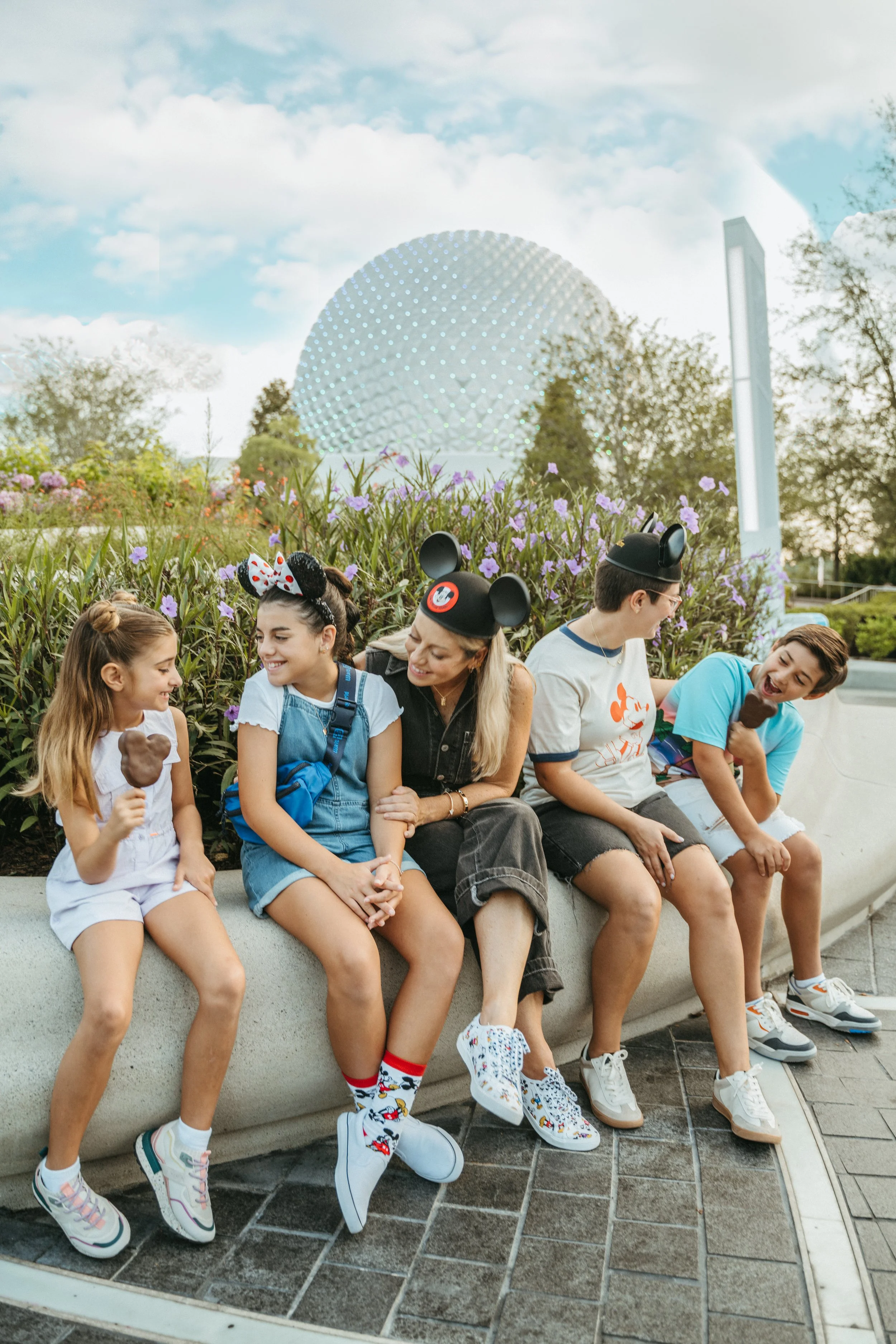 A group of six children sitting on a low concrete wall in front of purple flowers, with a large geodesic dome and a tall white building in the background. The children are smiling and wearing casual clothing, some with Mickey Mouse ears or themed socks.