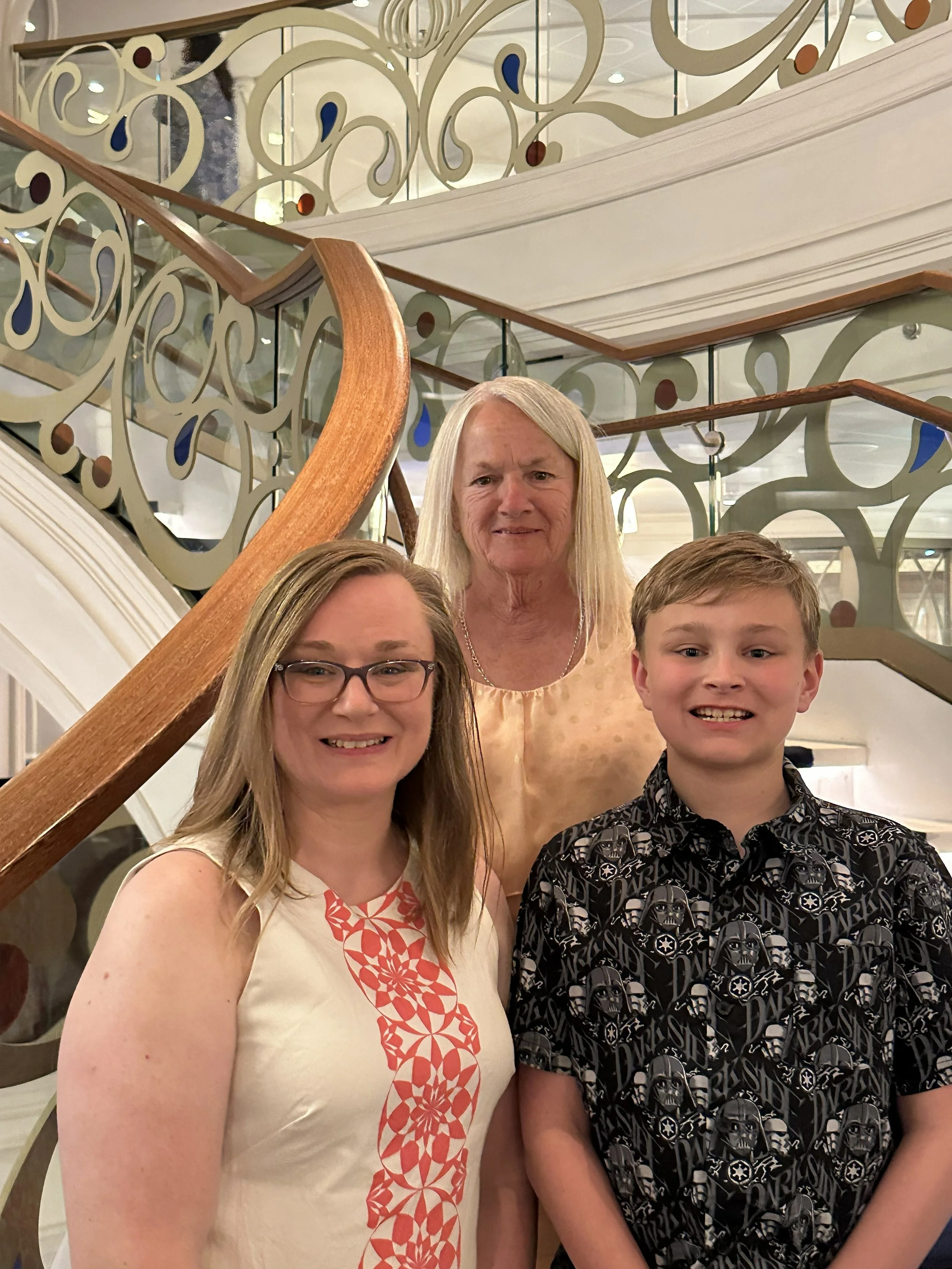 Group photo of three women and one young man standing on a staircase with ornate railing, smiling indoors.
