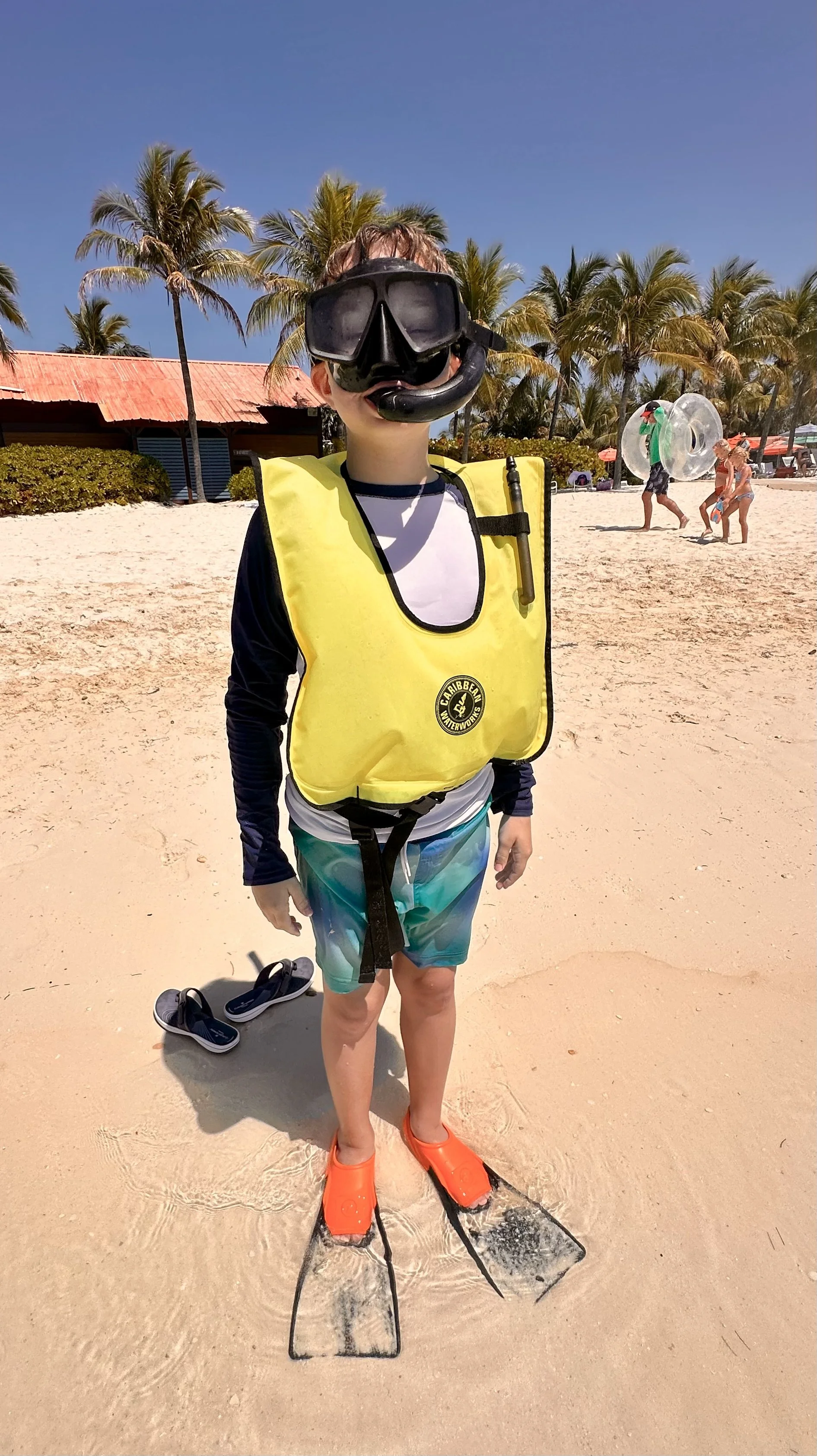 Child on a sandy beach wearing a yellow life jacket, snorkeling mask, and fins, with palm trees and beachgoers in the background.