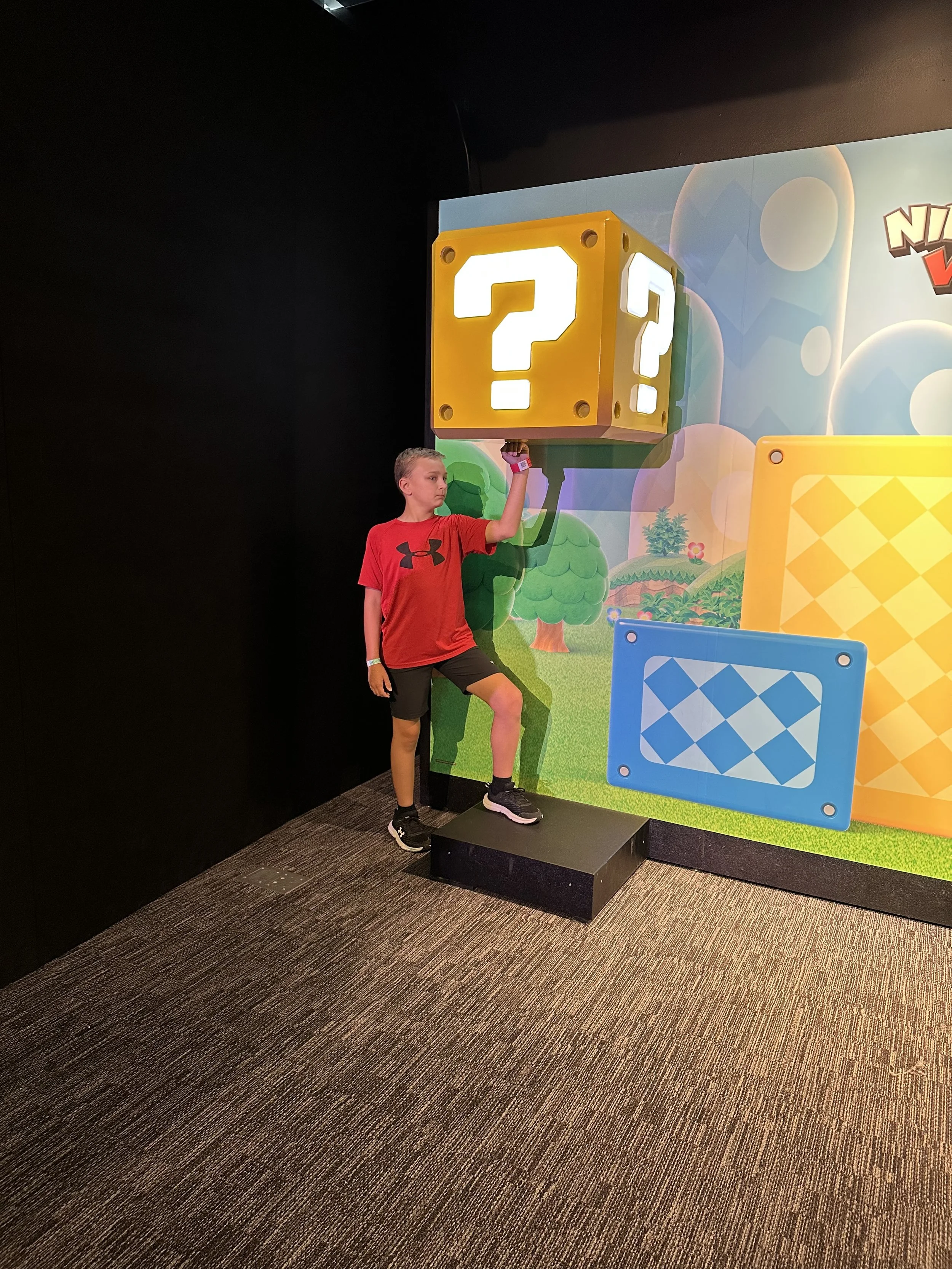 A young boy in a red t-shirt and black shorts standing on a black platform, reaching up with his right hand to touch a large illuminated question mark on a colorful Mario-themed backdrop.