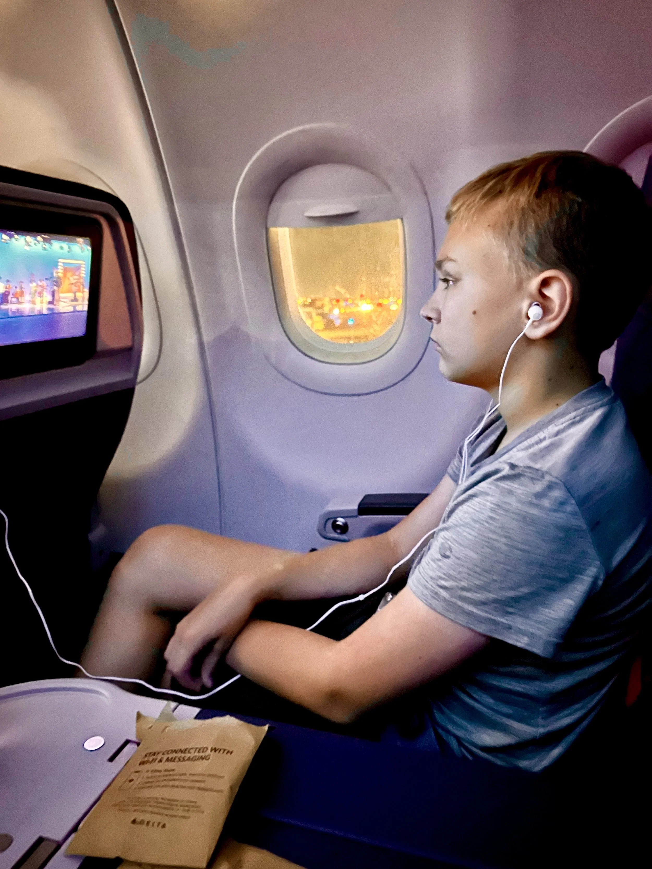 A young boy sitting in an airplane seat, watching a movie on the in-flight entertainment screen, with earphones in, and a window showing raindrops on the glass.