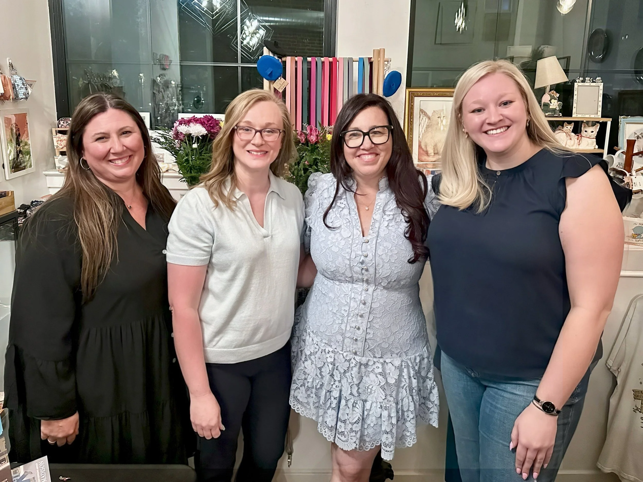 Four women smiling and standing together indoors, with shelves and decor in the background.