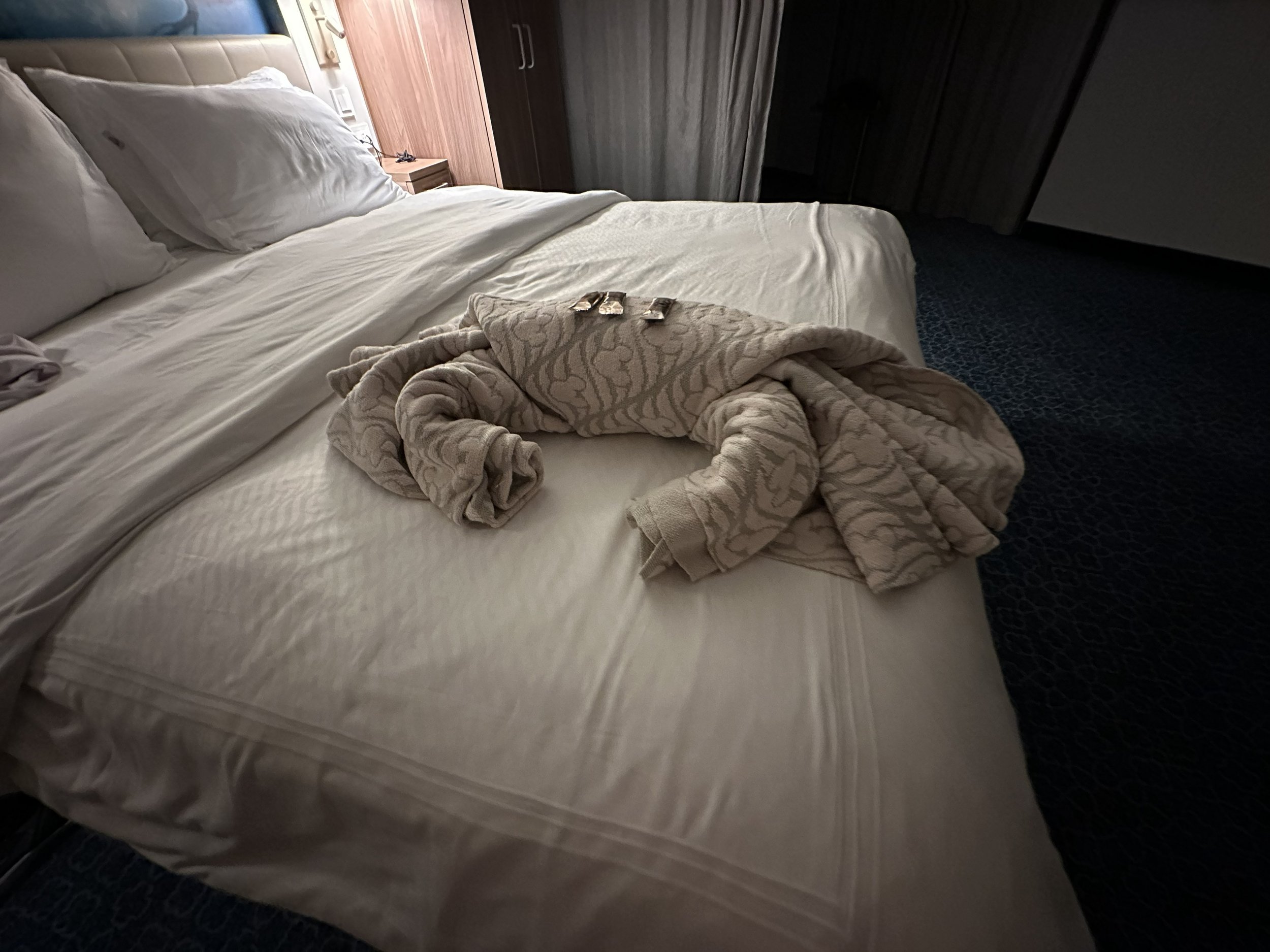 A neatly made bed with white bedding, pillows, and a beige blanket folded on top. A towel art in the shape of a butterfly is placed on the bed. There is a wooden headboard, a small nightstand, and a wardrobe in the background.