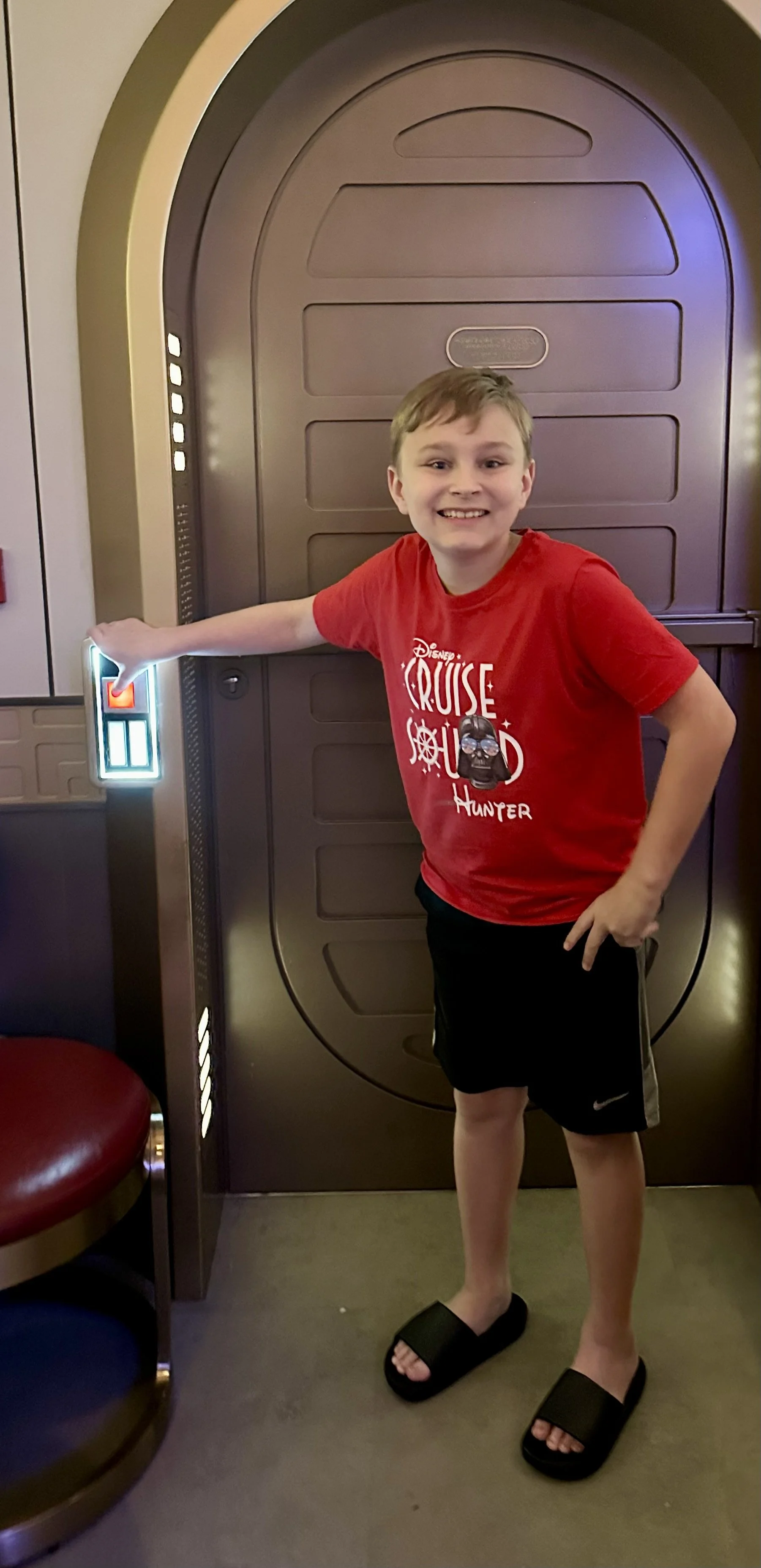 A boy in a red Disney Cruise Line shirt and black shorts standing in front of a spaceship door, smiling with one hand on a control panel.