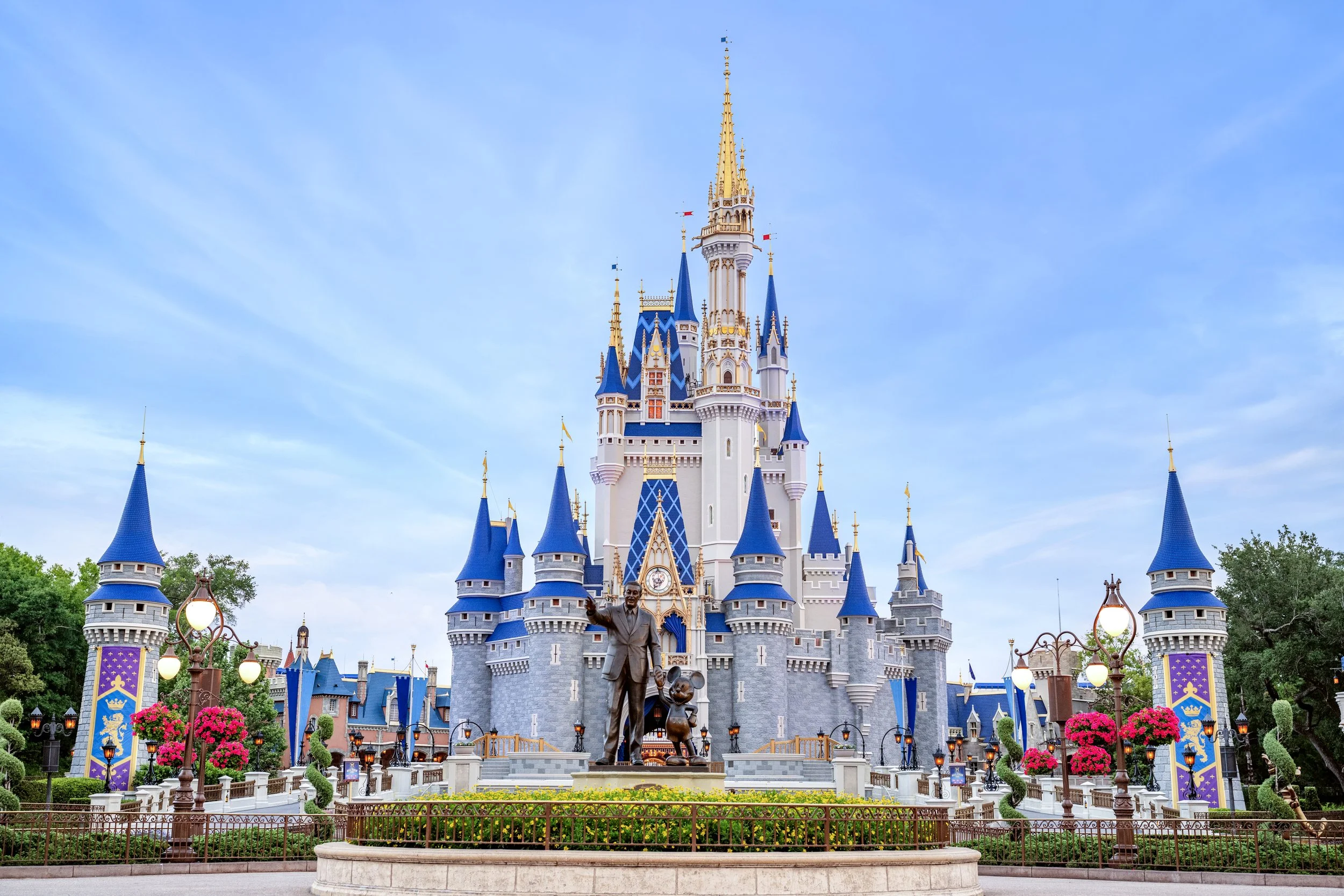 A castle replica at Disneyland with blue spires, gold accents, and a statue of Walt Disney and Mickey Mouse in front, surrounded by landscaped gardens and pink flowers.