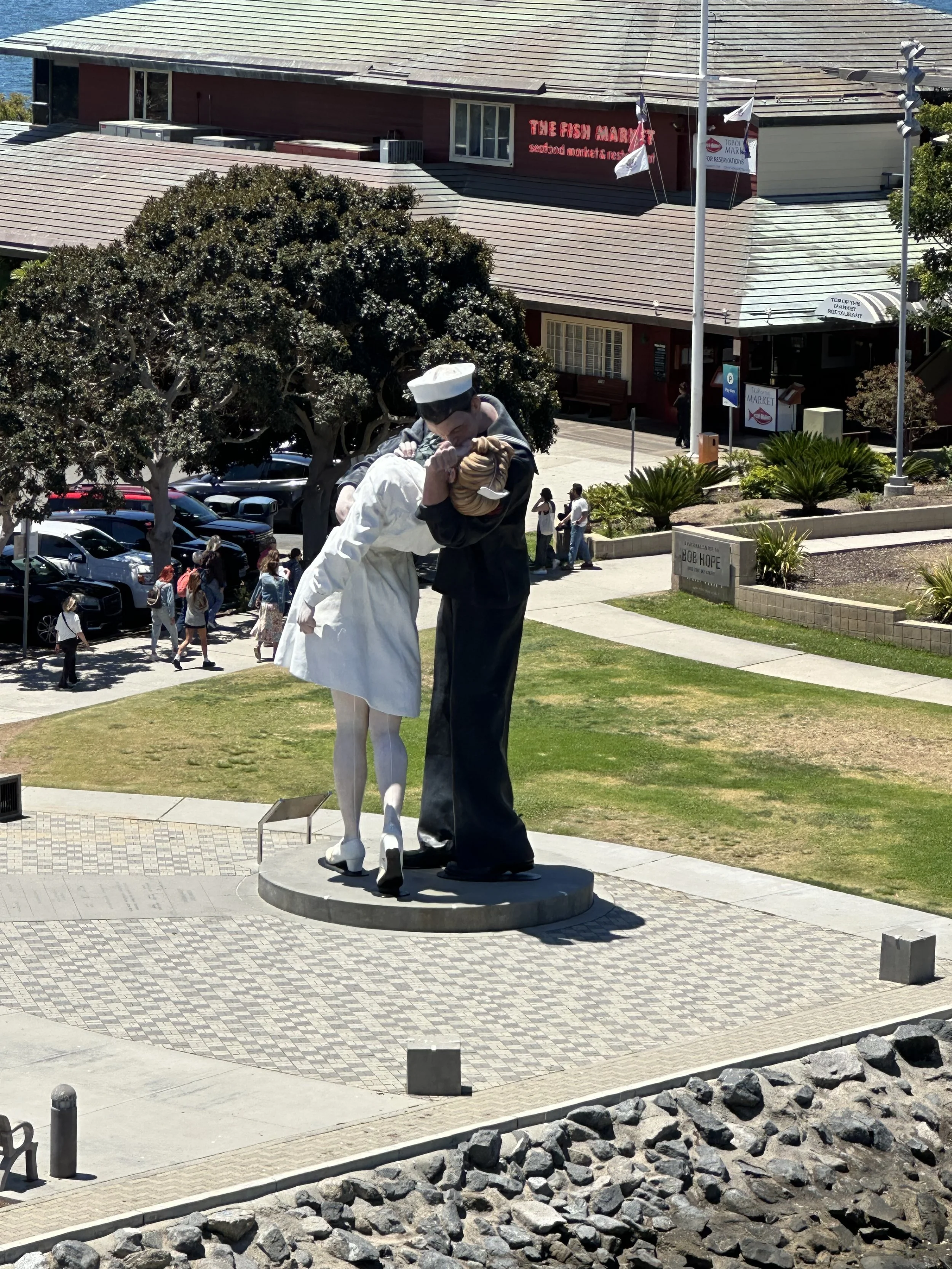 A sculpture of a sailor kissing a nurse stands on a platform in an outdoor area, with trees, cars, and buildings in the background. In San Diego, California from the Midway. 