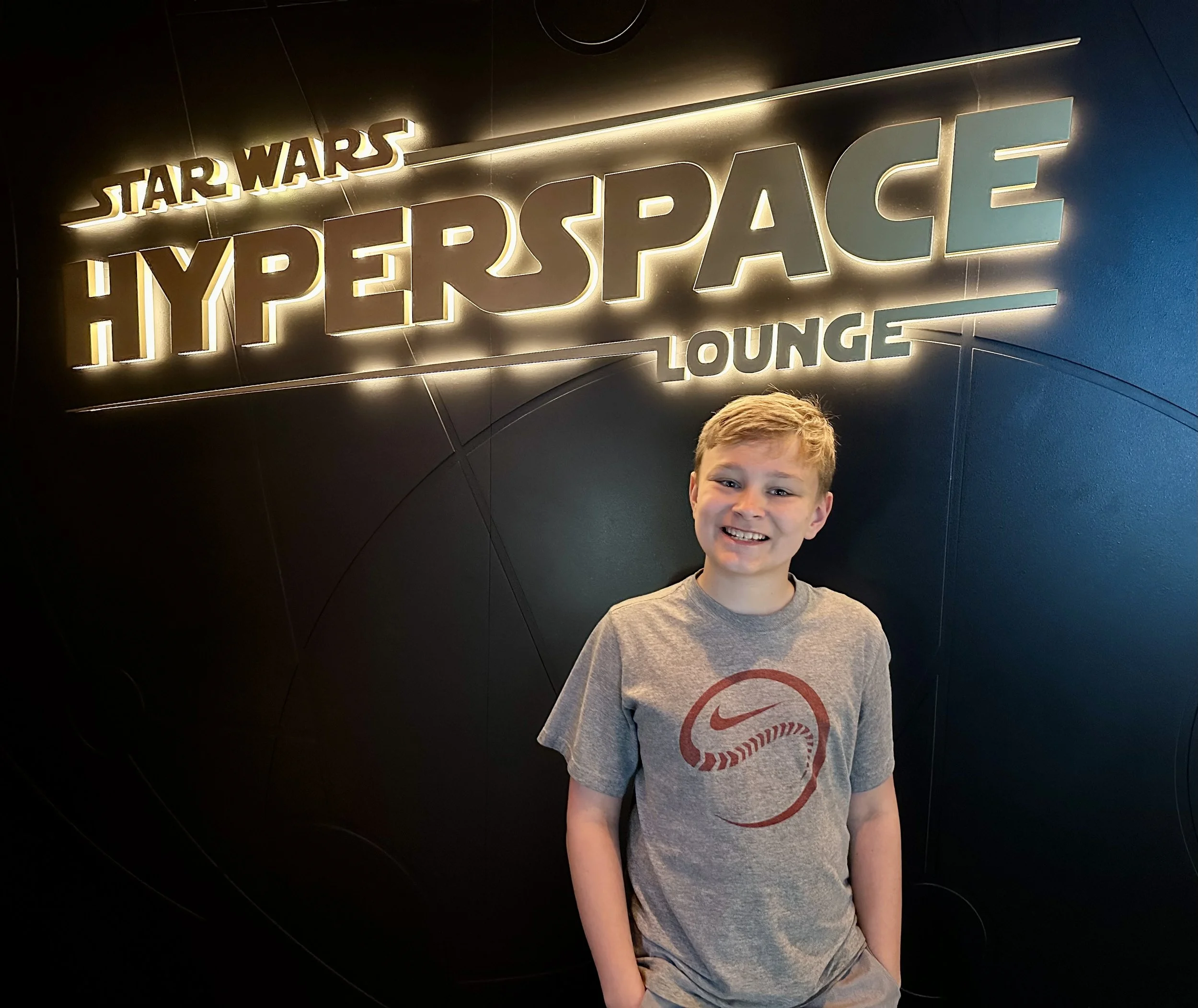 A smiling boy with short hair standing in front of a black wall with illuminated sign that reads 'Star Wars HyperSpace Lounge'.