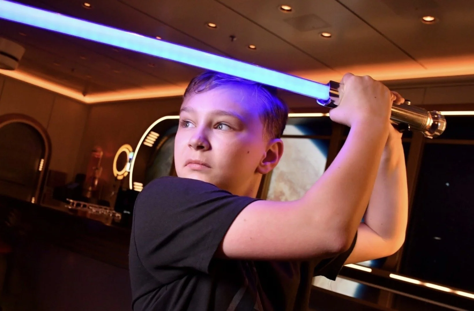 A young boy with short hair holding a glowing blue lightsaber in a room with futuristic and space-themed decor.