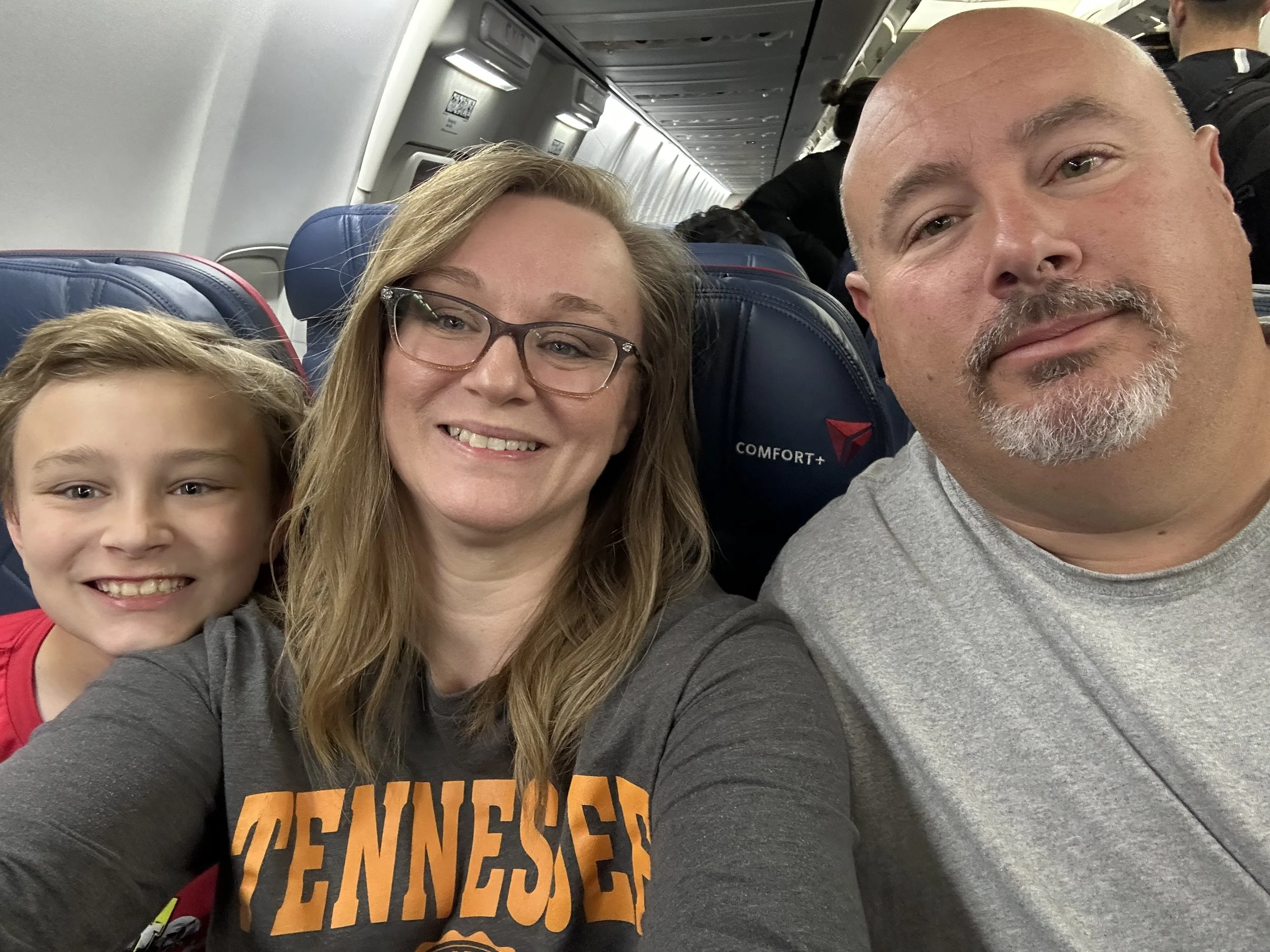 A family of three taking a selfie on an airplane. The woman and child are smiling, and the man is looking at the camera with a neutral expression.