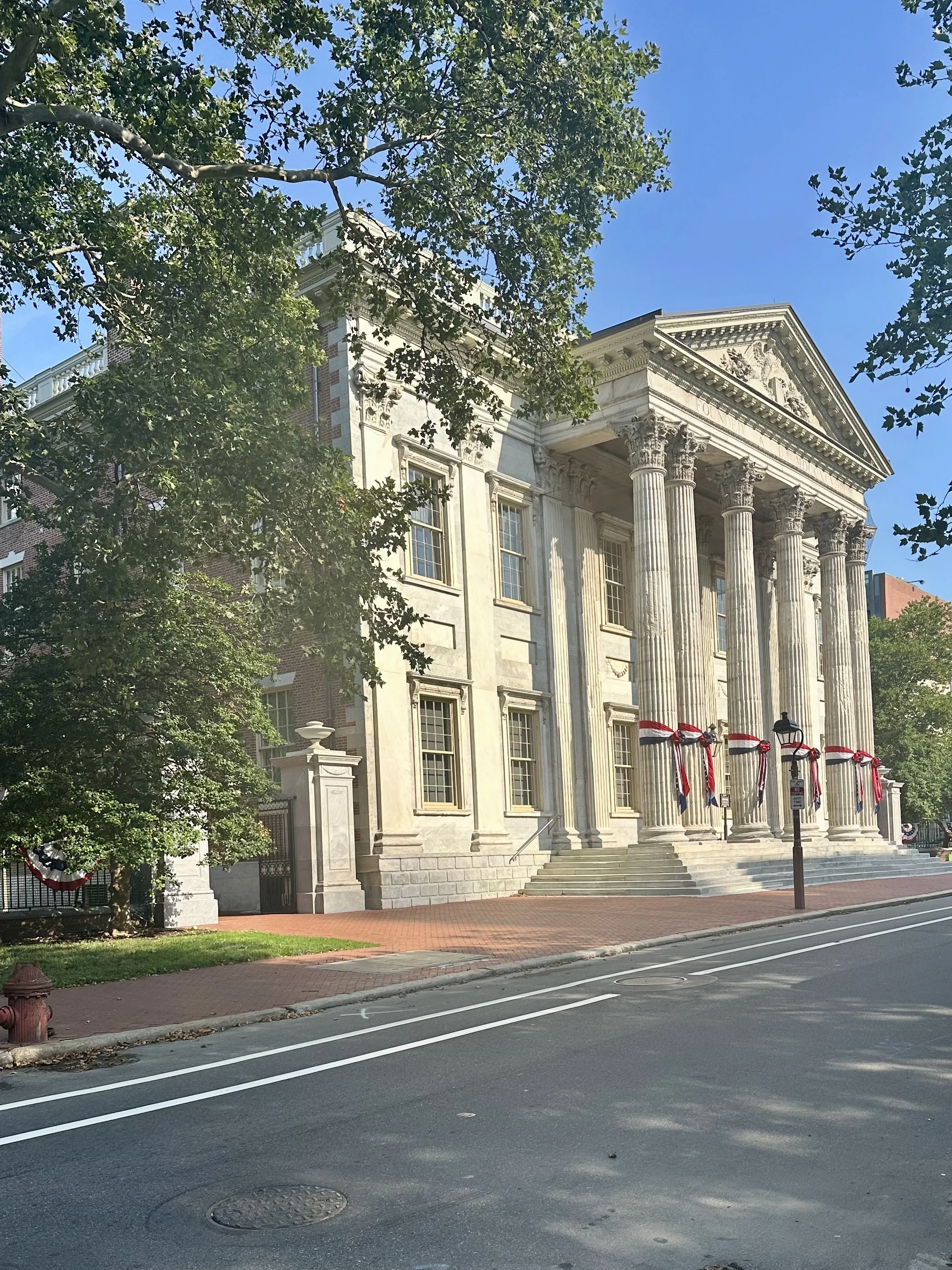 Historical building with classical columns and flags decorated with red, white, and blue ribbons, trees in the foreground, and a street in the foreground.