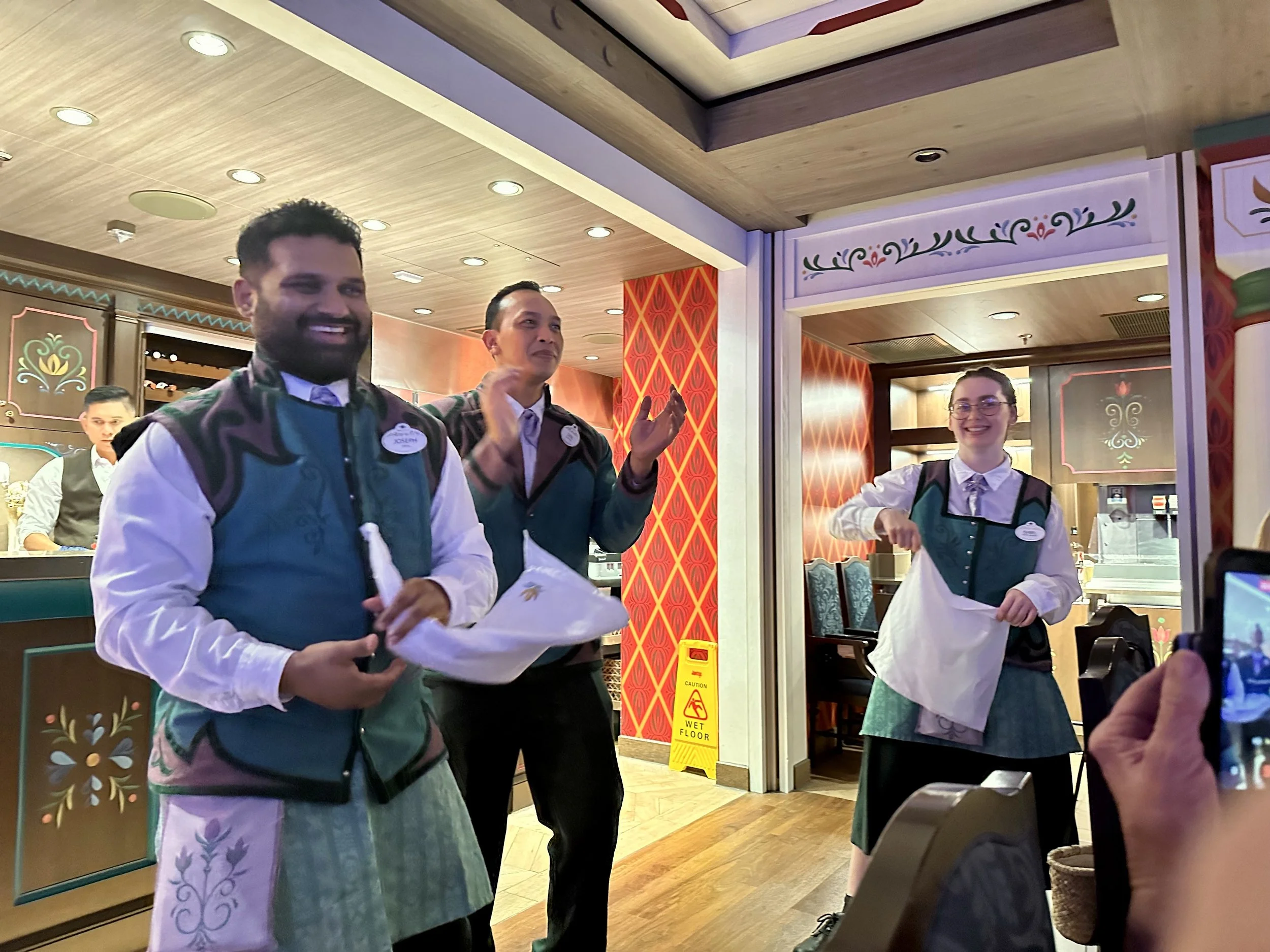 Three Disney employees, two men and a woman, smiling and clapping behind a counter inside a Disney restaurant. The staff are dressed in colorful uniforms with aprons and nametags, and the background features decorative wall panels and a sign warning 