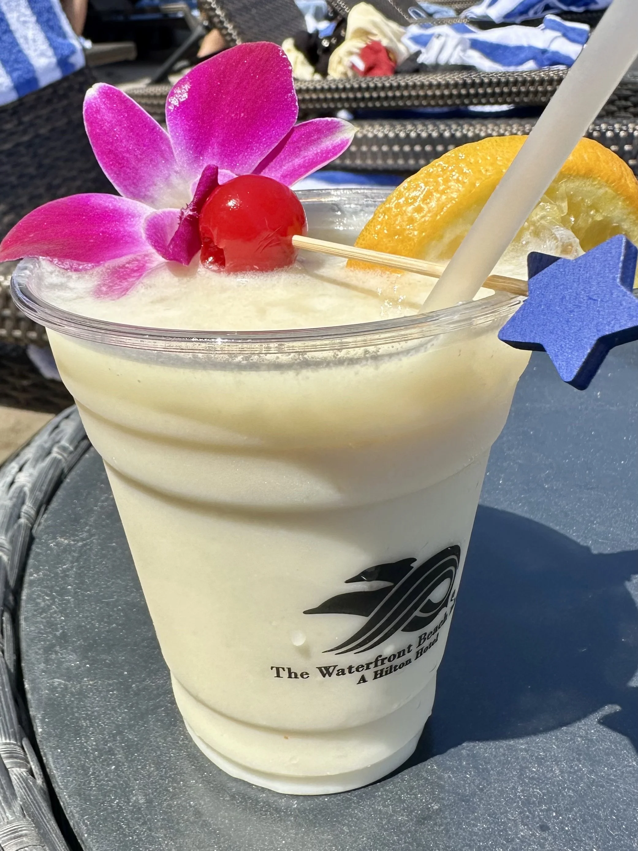 A tropical cocktail in a clear cup decorated with a pink orchid flower, a red cherry on a skewer, a slice of lemon, and a straw, sitting on a metal table outside.