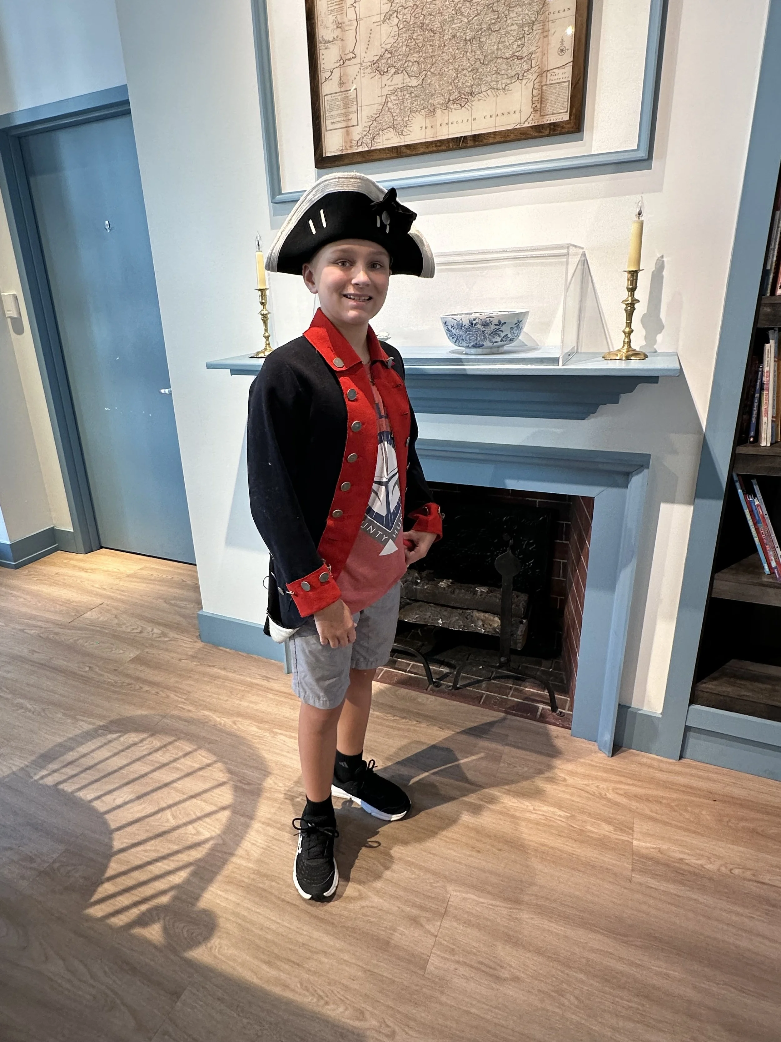 A young boy standing in front of a fireplace with a nautical-themed costume, wearing a pirate hat, a black and red jacket, and gray shorts. There are candleholders and a bowl on the mantel, with a framed map hanging above.