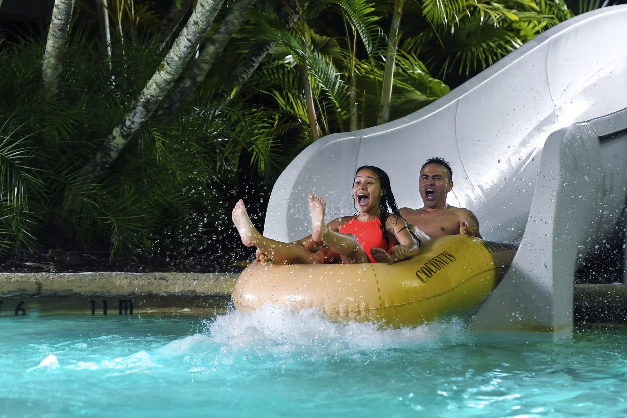 Two people riding a water slide into a pool, surrounded by lush tropical plants.