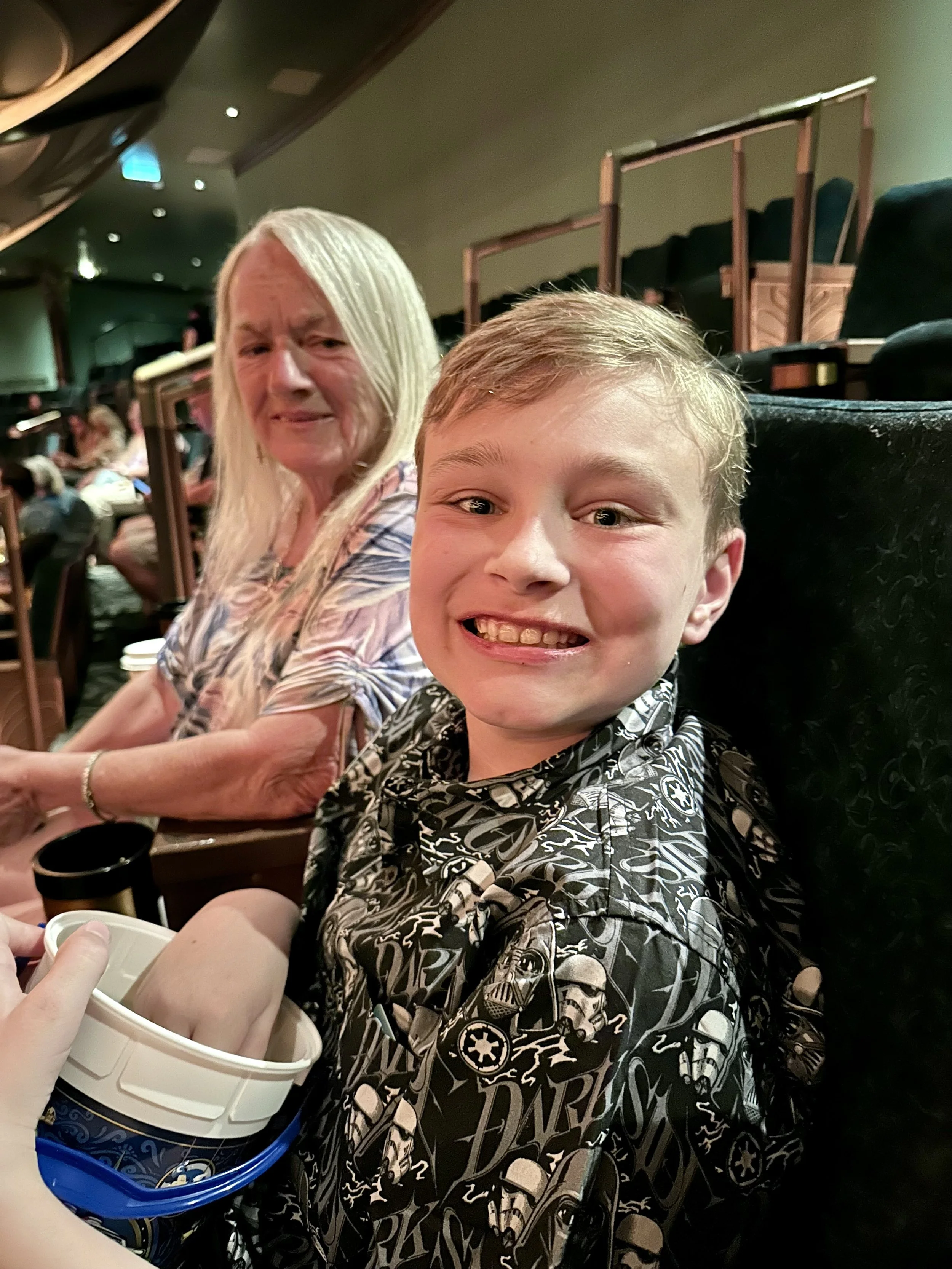 A boy smiling excitedly at the camera with a woman sitting behind him at a restaurant, with a bucket of food in the foreground and other diners in the background.