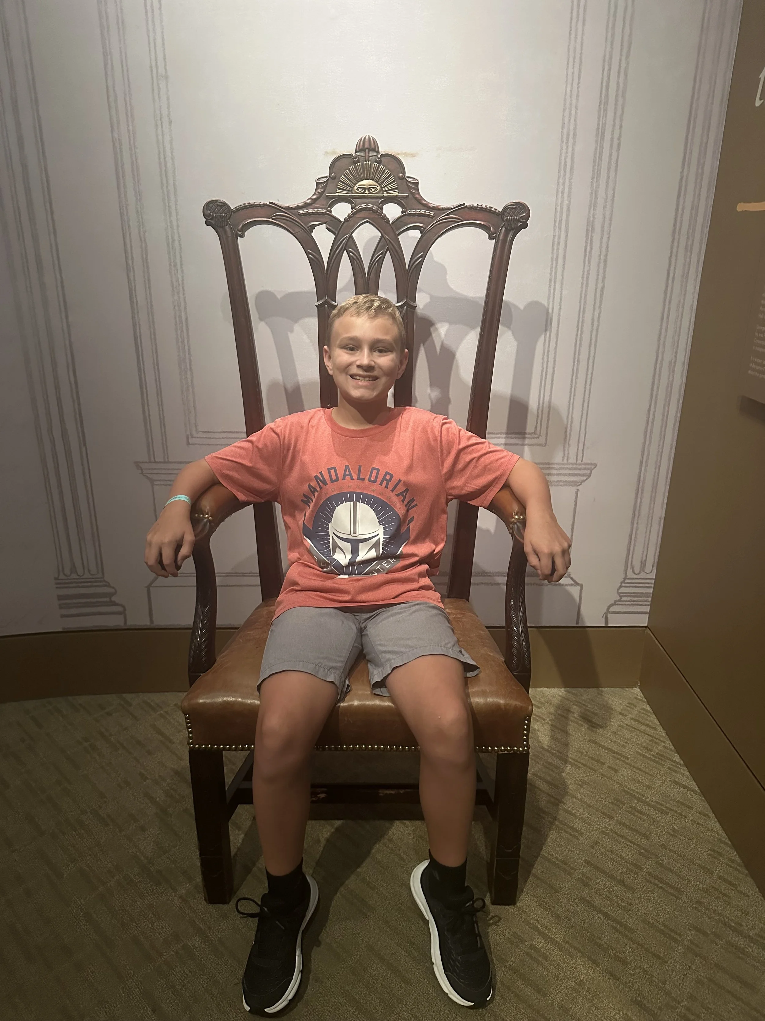 A young boy with short light hair is sitting on a tall wooden chair with a high backrest and ornate design, against a background with a decorative wall. He is smiling, wearing a pink T-shirt, gray shorts, black sneakers, and a wristband on his left w