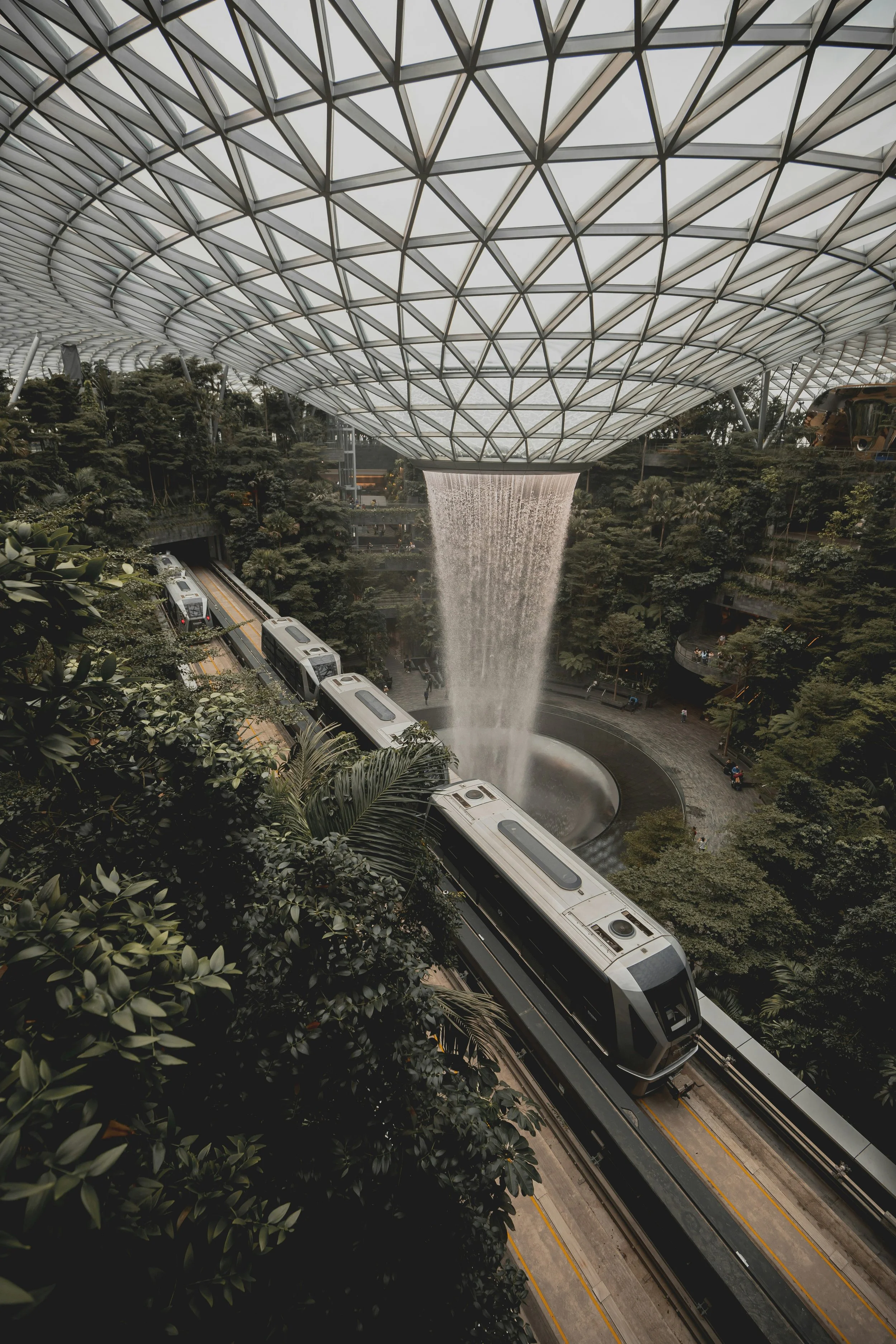 Indoor garden with a waterfall feature, a glass and metal roof, trees, and a train on an elevated track.