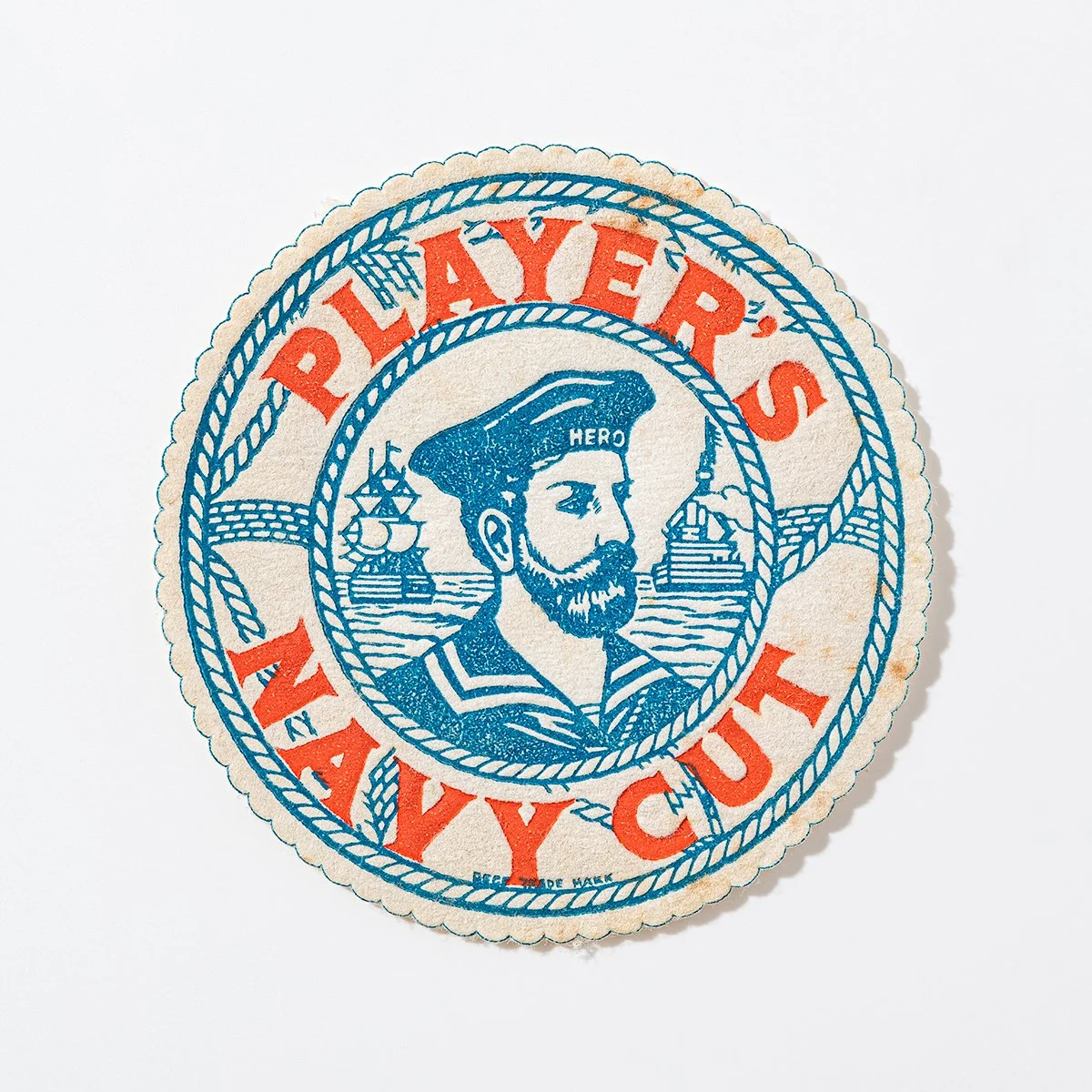 A circular sticker or badge with a nautical design. It features an illustration of a sailor with a beard and a hat labeled 'HERO' in the center. Around the sailor, there are ships and sea waves. The outer text reads 'PLAYER'S NAVY CUT' in red, with a blue rope design encircling the inner illustration.