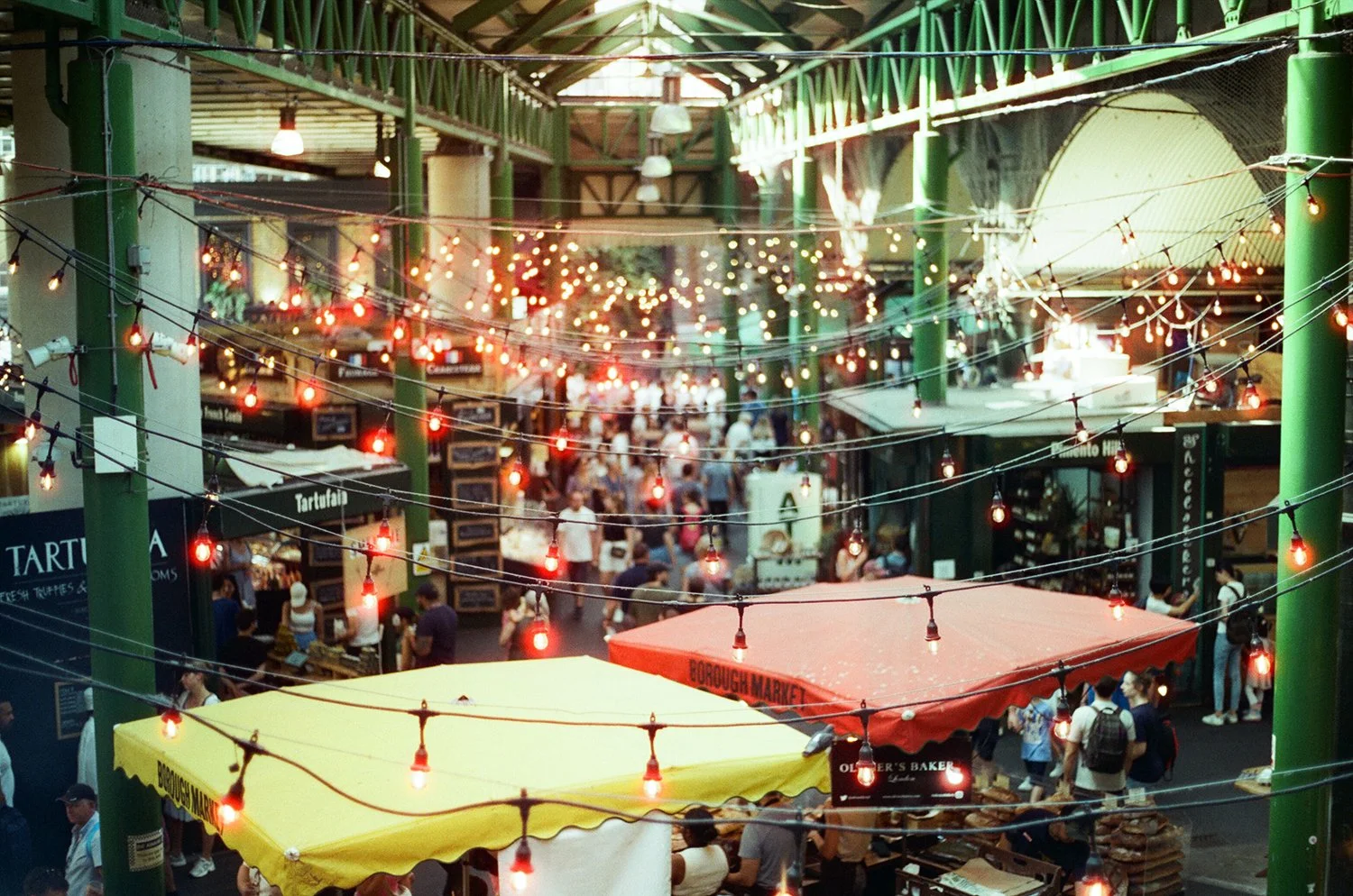 Indoor marketplace with string lights hanging overhead, yellow and red market stalls, and numerous shoppers walking among vendor booths.