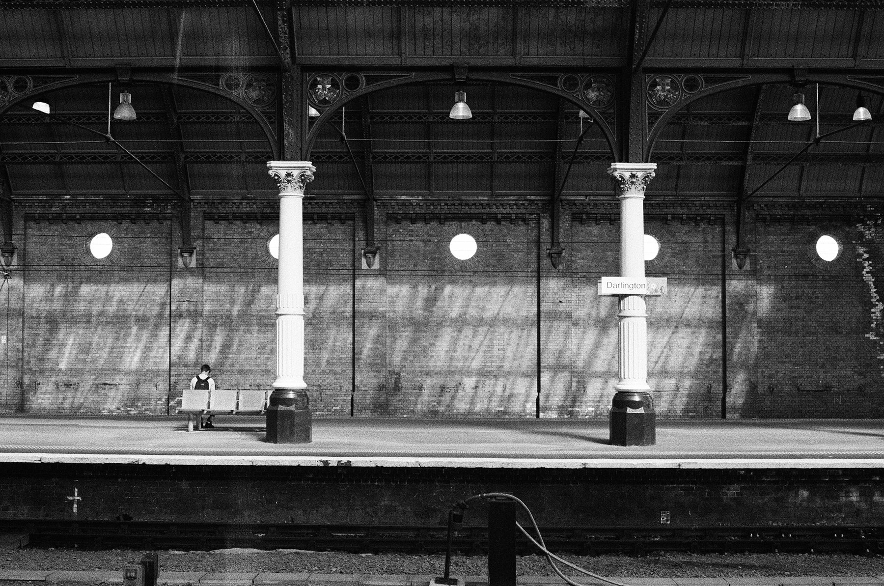 A black and white photo of a train station platform with a brick wall and ornate columns. There is a woman sitting on a bench at the far left. The platform has a sign that reads 'Darlington'.