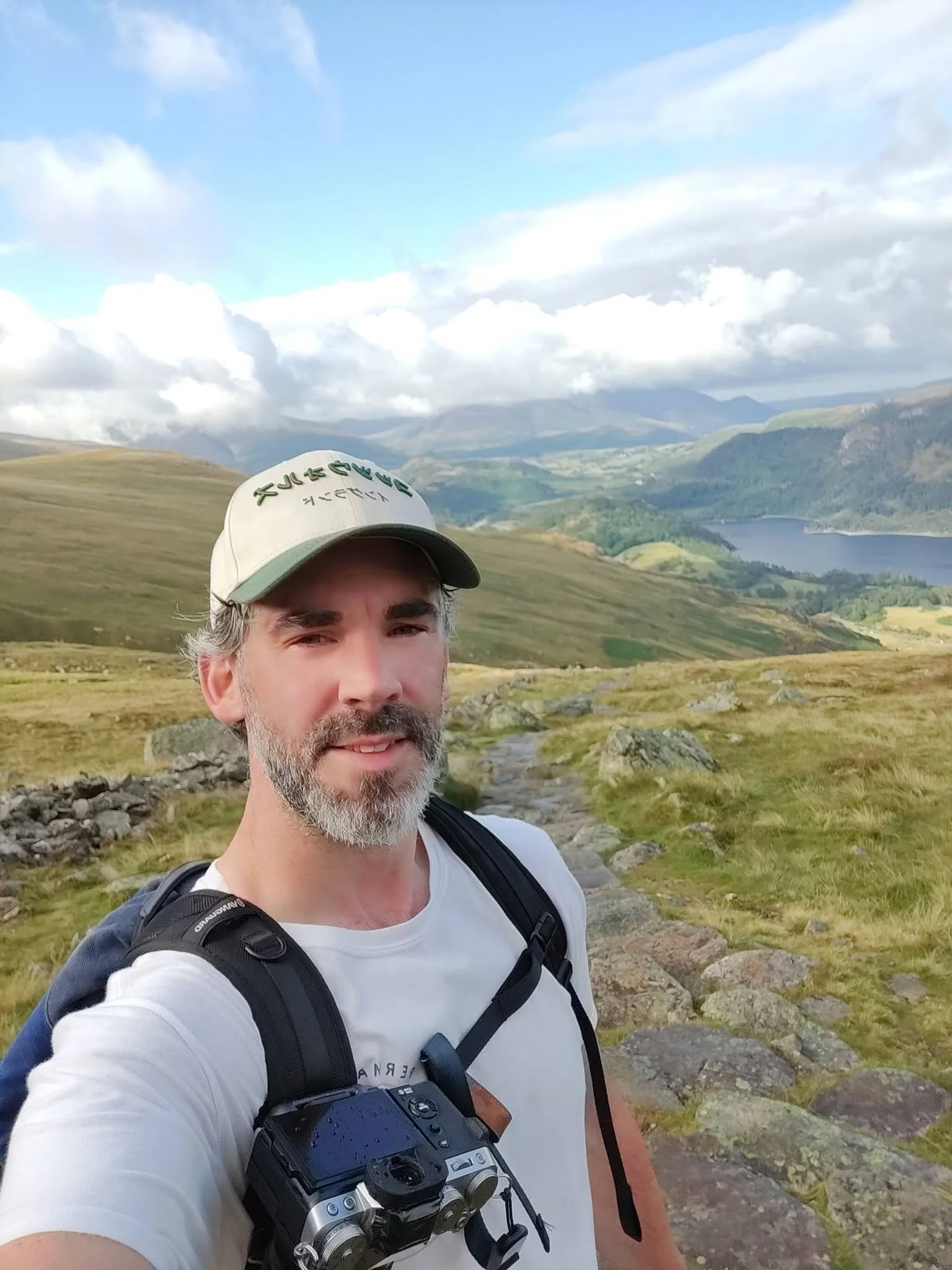 A man with a gray beard, wearing a cap, white t-shirt, and a black backpack, taking a selfie in a mountainous landscape with green valleys, a river, and cloudy sky.