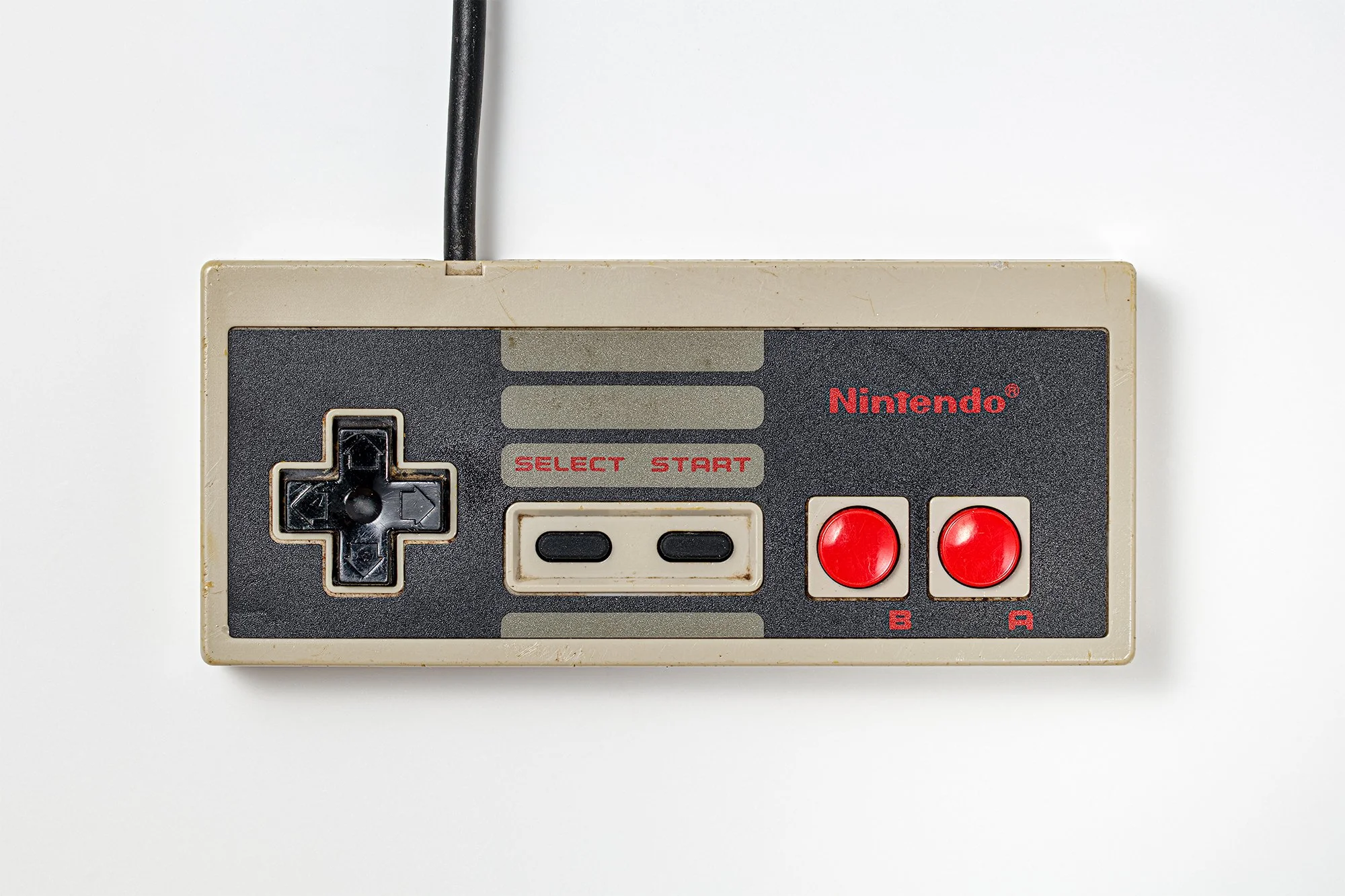 A vintage Nintendo gaming console with beige and black colors, featuring a directional pad, 'SELECT' and 'START' buttons, and red 'A' and 'B' buttons, connected to a black wire.