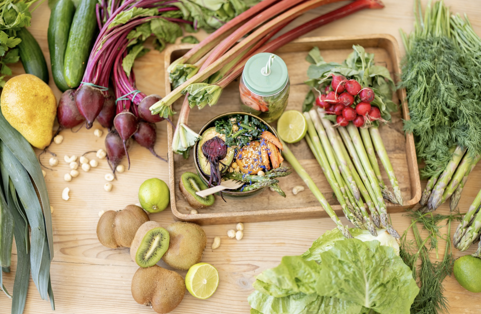 An assortment of fresh vegetables, fruits, and a smoothie on a wooden table. Items include cucumbers, beets, radishes, asparagus, kiwi, lemon, lettuce, and a jar of green juice.