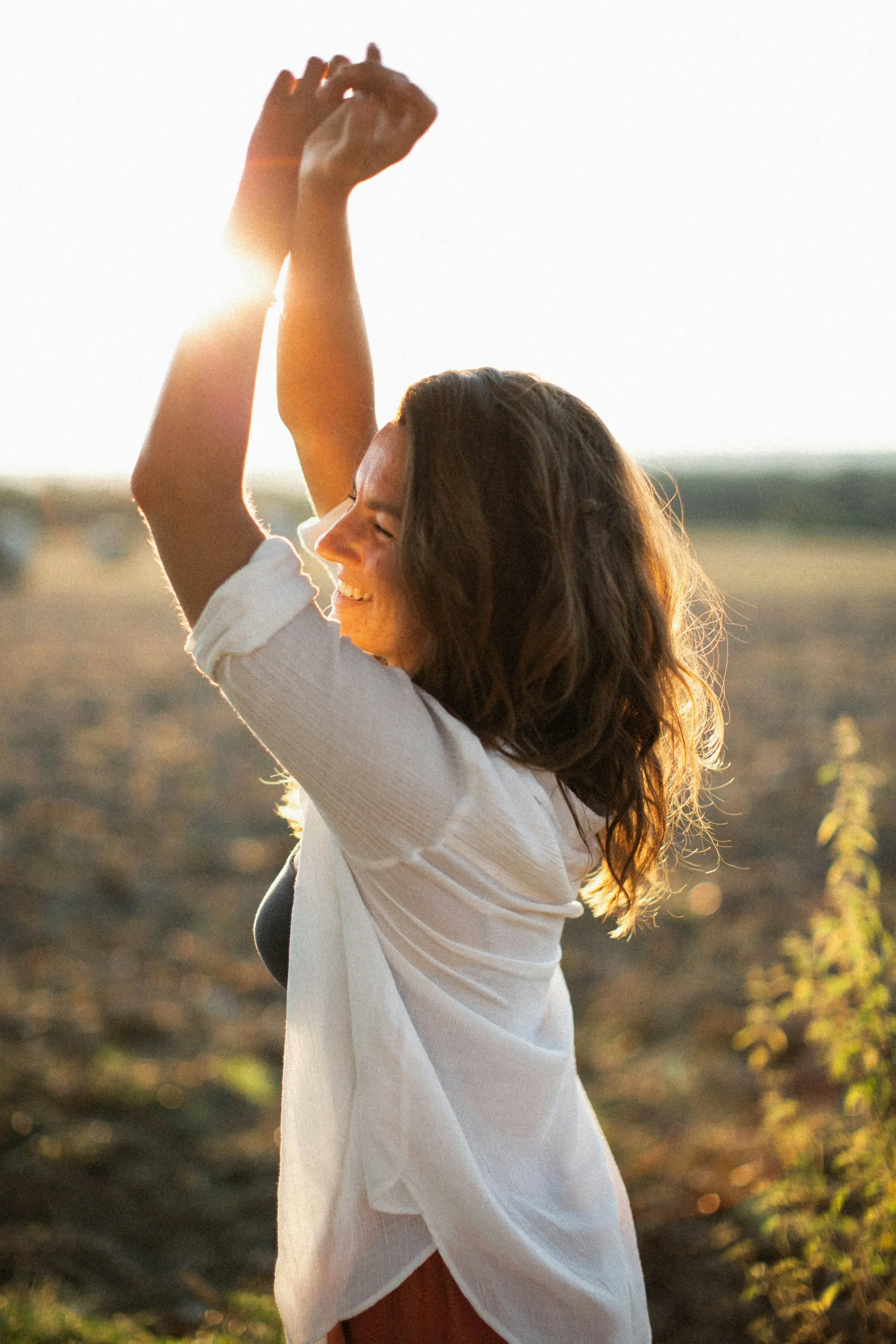 A woman with long dark hair smiling and raising her arms in a field during sunset.