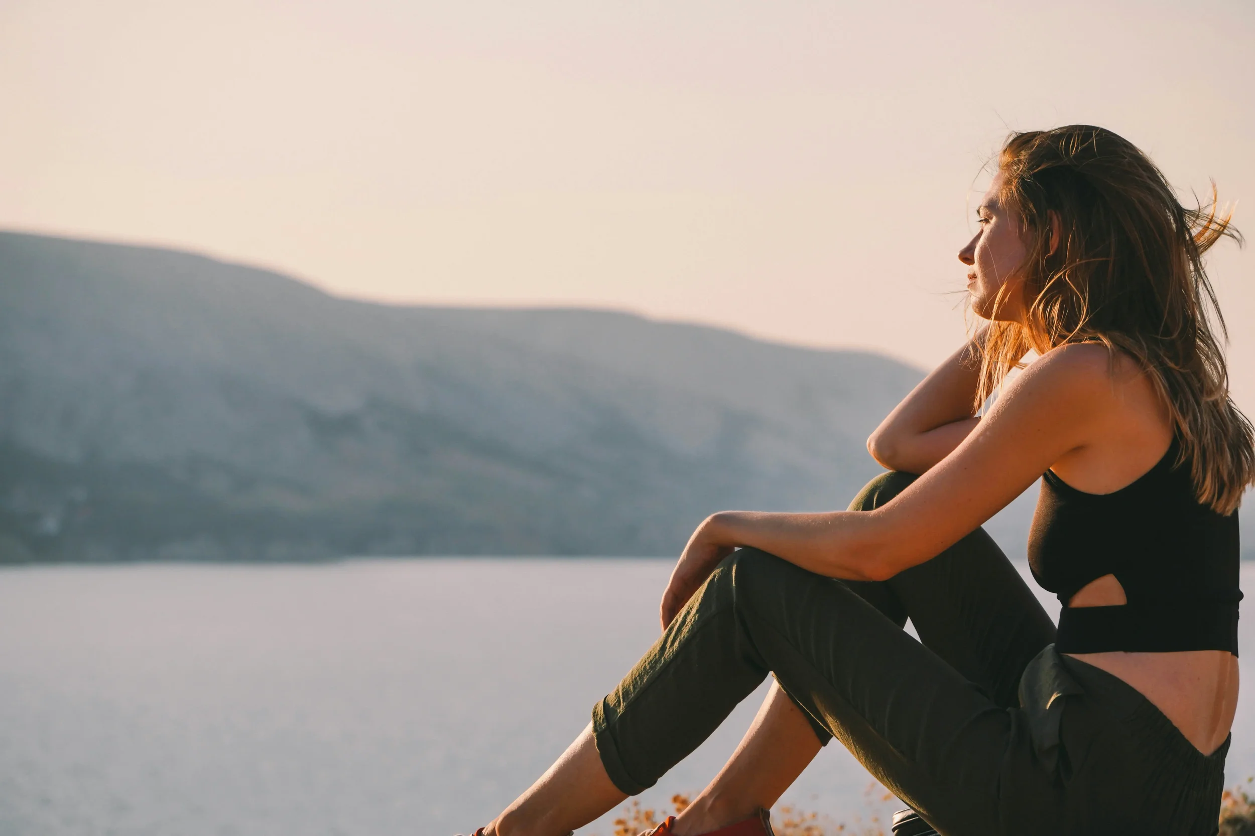 A young woman with long hair sitting outdoors during sunset, watching the horizon with mountains in the background.