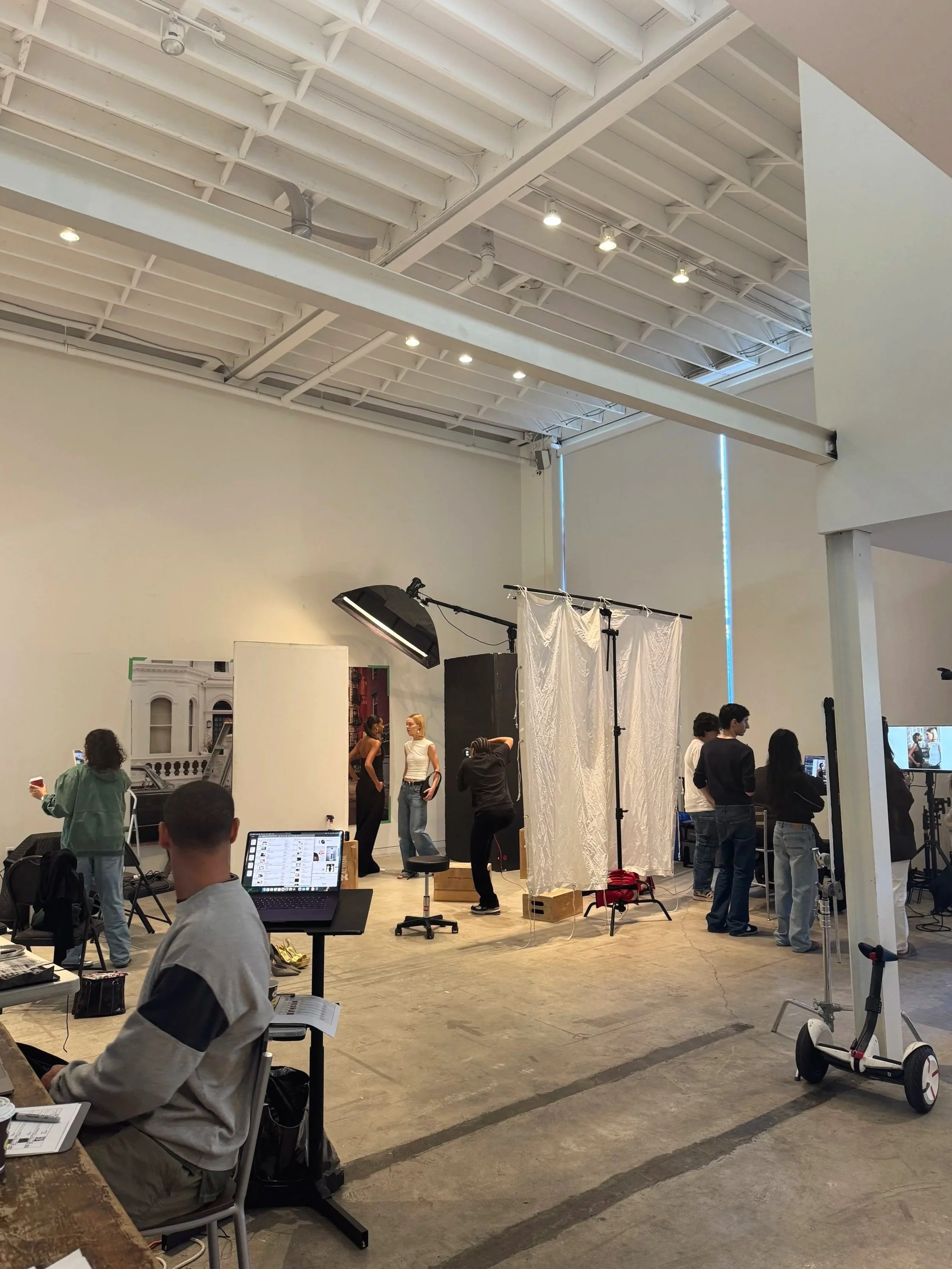Behind the scenes of a fashion and beauty photoshoot. Models pose against a city backdrop, with v-flats, photography light, silk, and production crew in the frame.