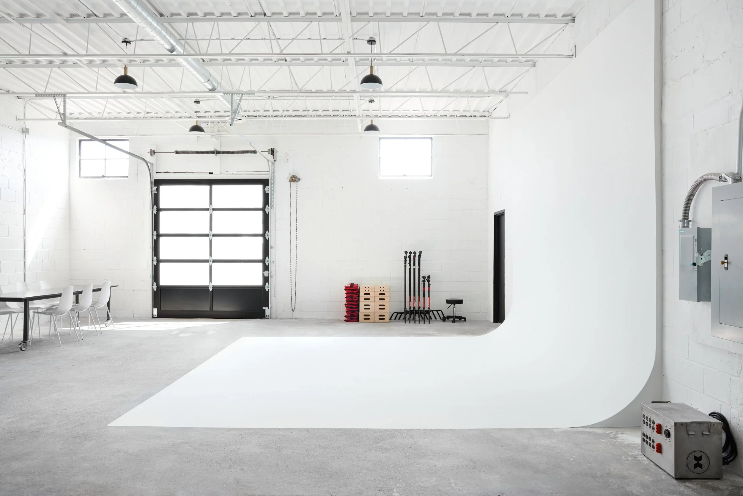 16' x 12' x 15' white cove. Studio shows a garage door, dining table, apple boxes, sandbags, c-stands, and electrical safety switch.