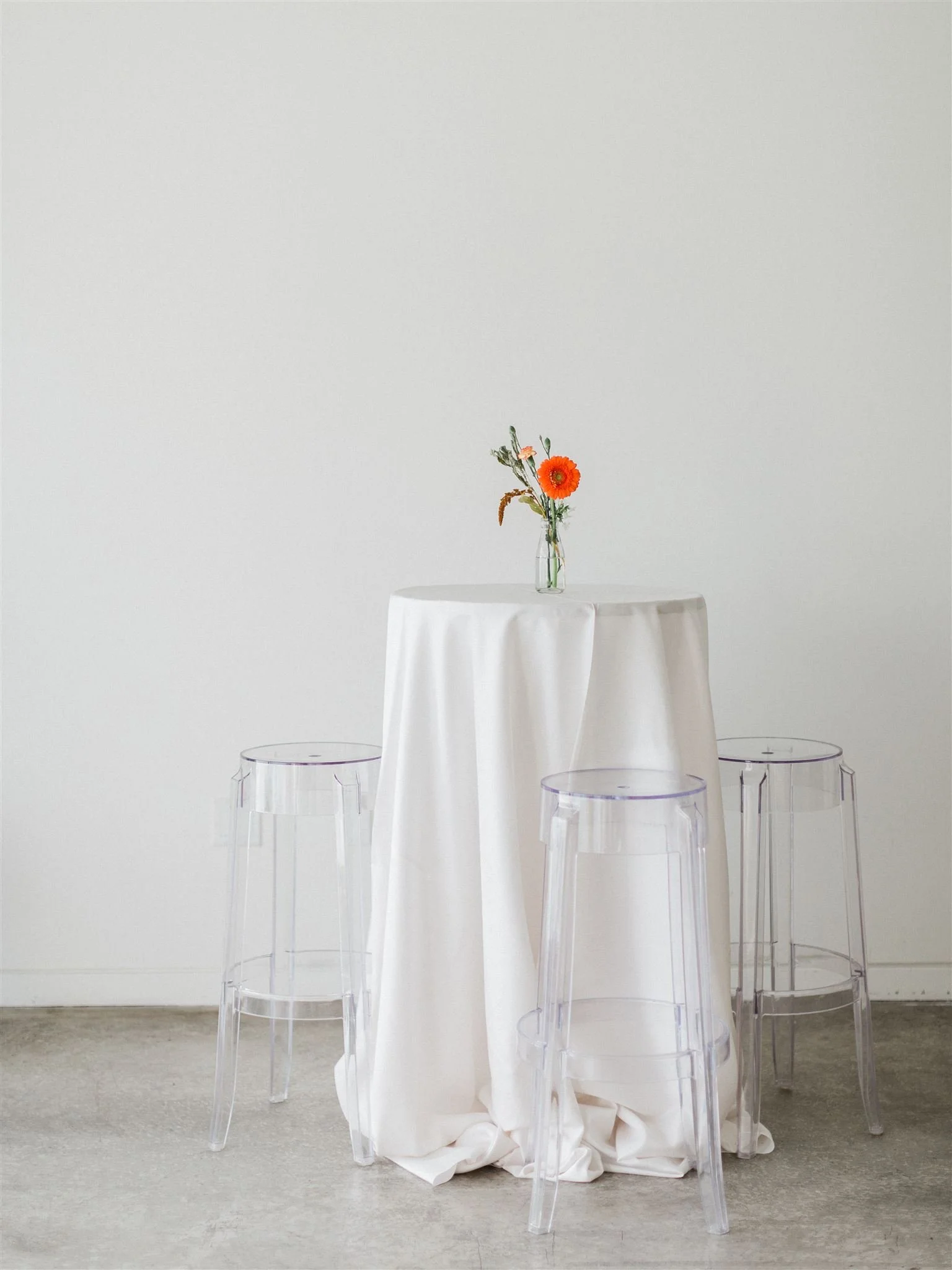 Three acrylic chairs, and a cocktail table covered with a long, classy tablecloth are placed inside a neutral and timeless Toronto event venue and studio space. The table has a bud vase and orange flowers as a wedding centrepiece.