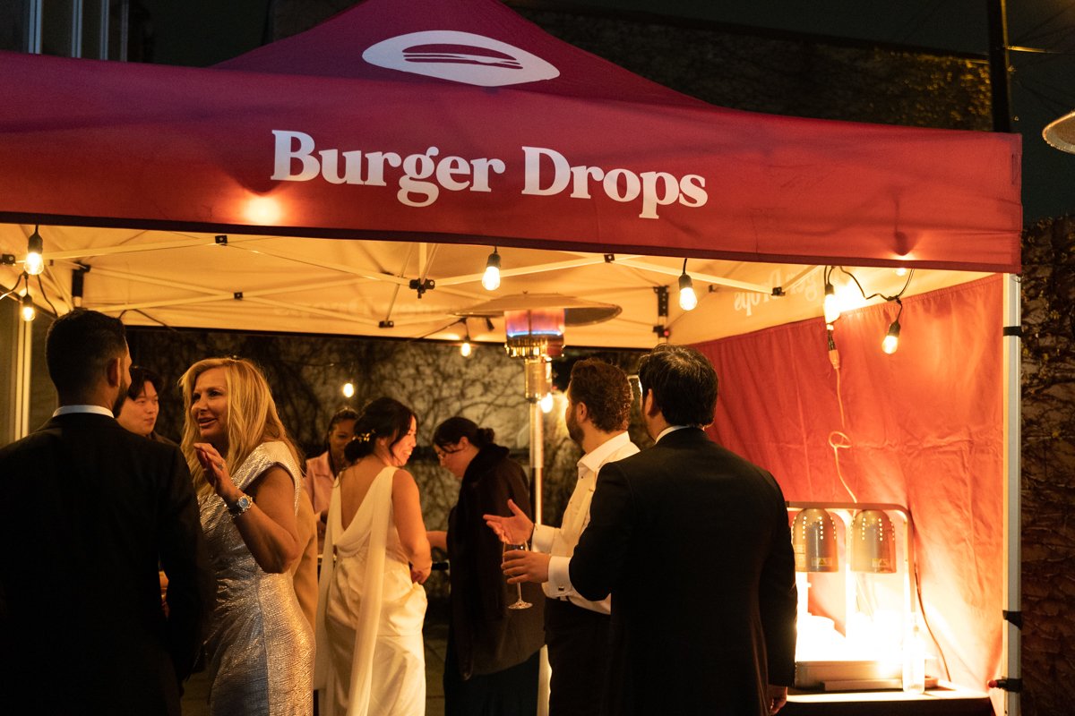 Wedding guests, bride, and groom chatting underneath Burger Drops tent lit by string lights 