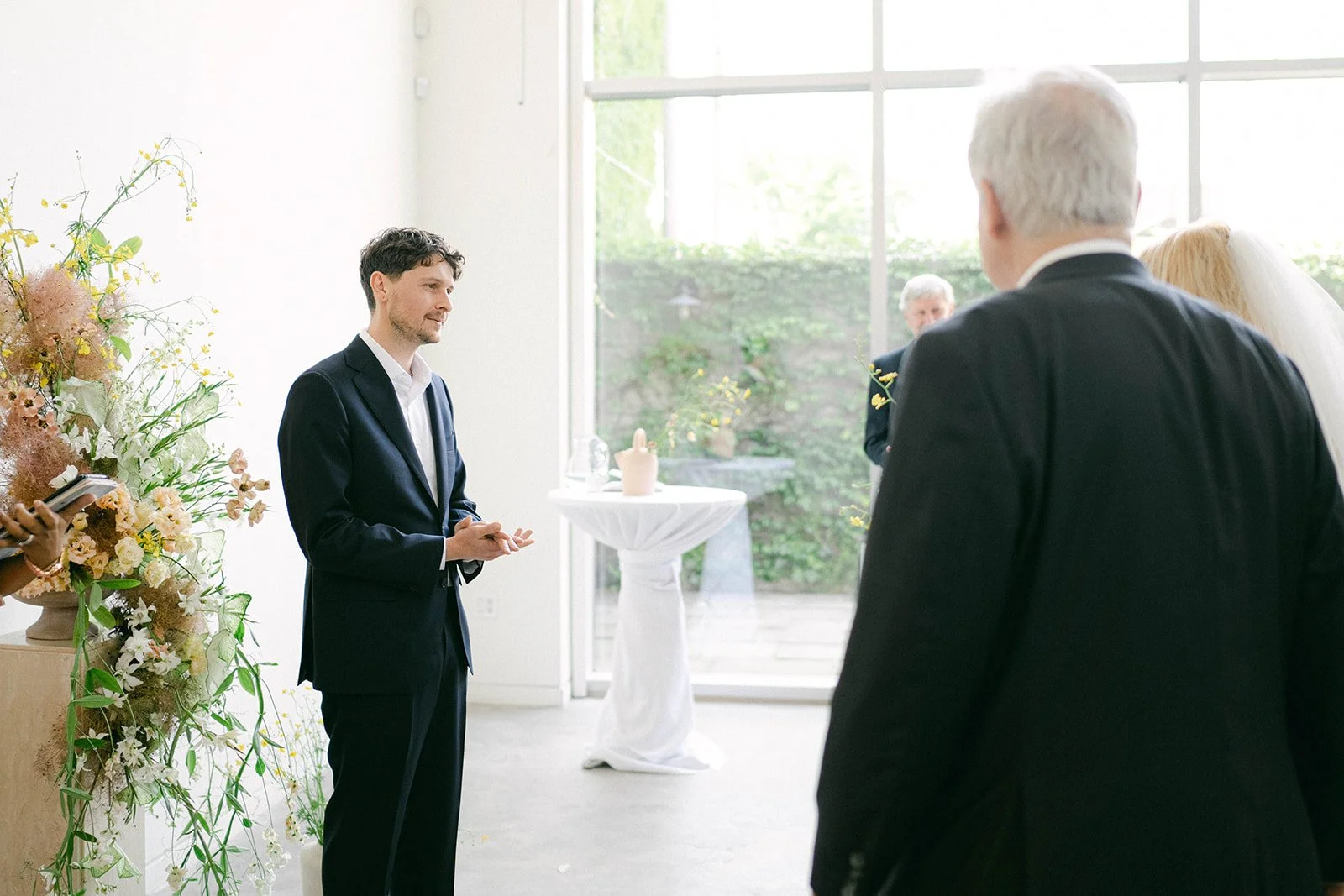 Groom looks at bride walking down the aisle inside a Toronto studio space and event venue with floor to ceiling windows and bright natural light