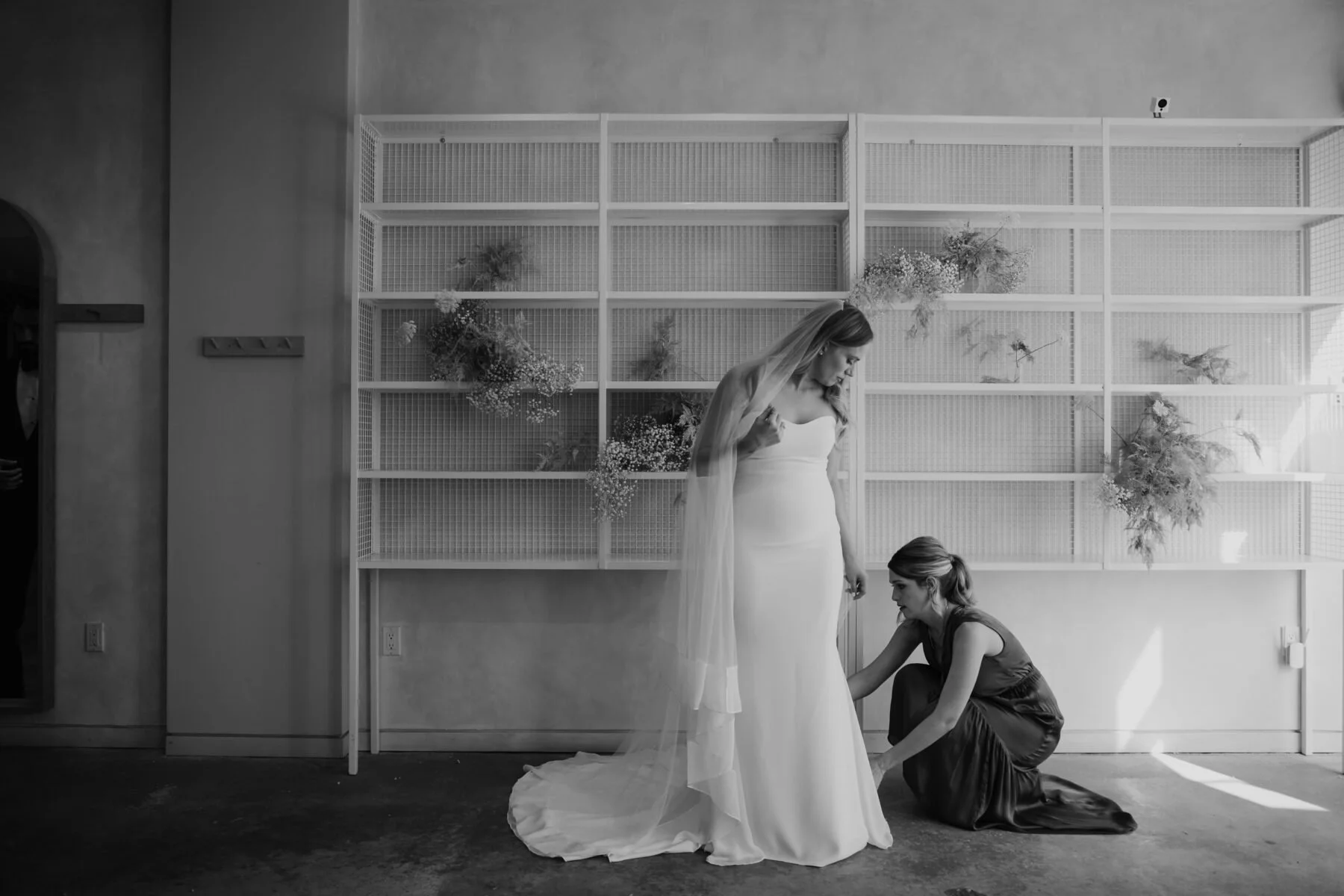 Studio lobby shelves decorated with florals. Bride stands in the foreground getting her wedding dress straightened out by a bridesmaid 
