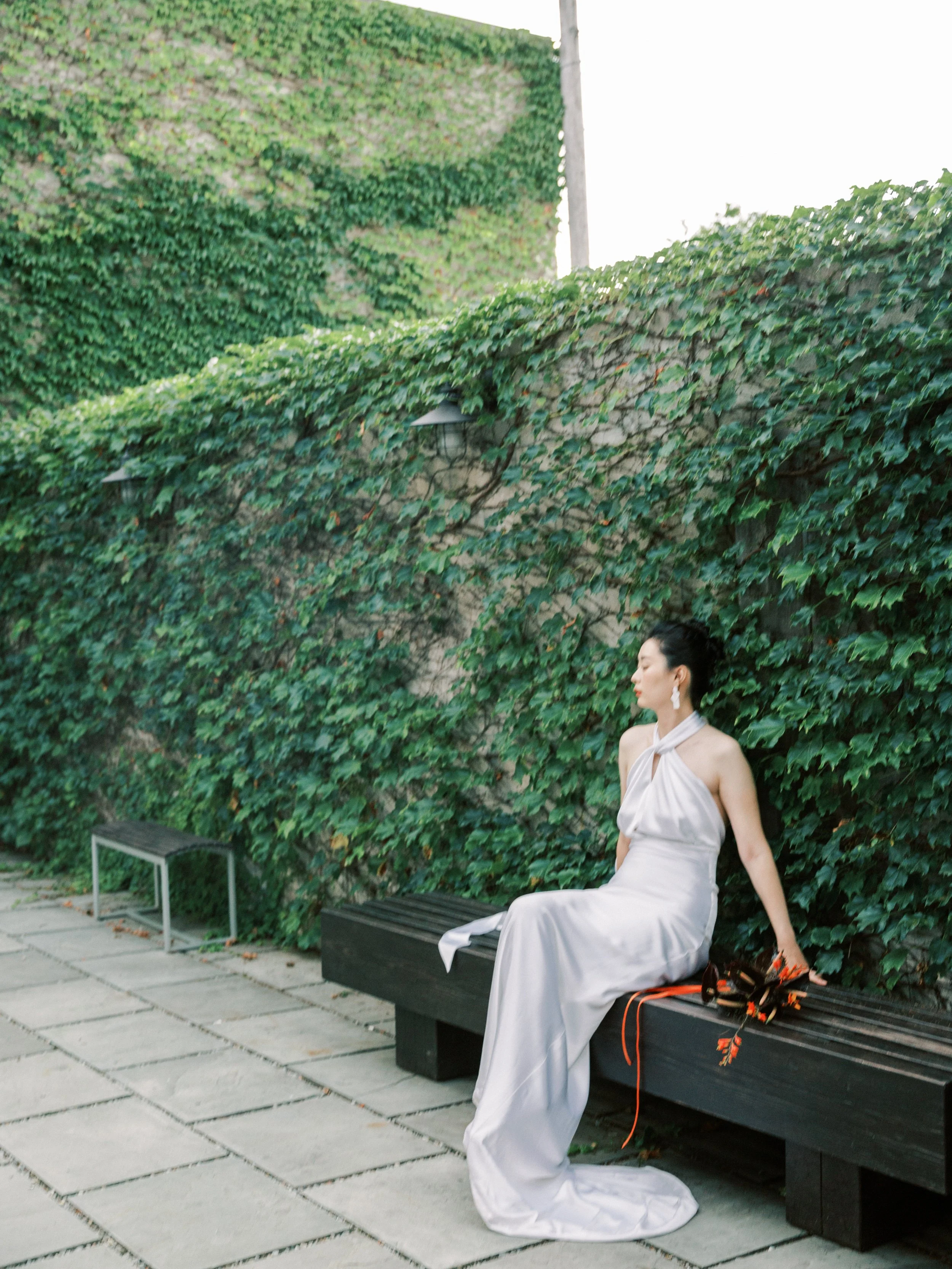 A bride sits outside a private patio bench with a wedding bouquet placed next to her. She is also learning against a tall vine and living green wall in the background.