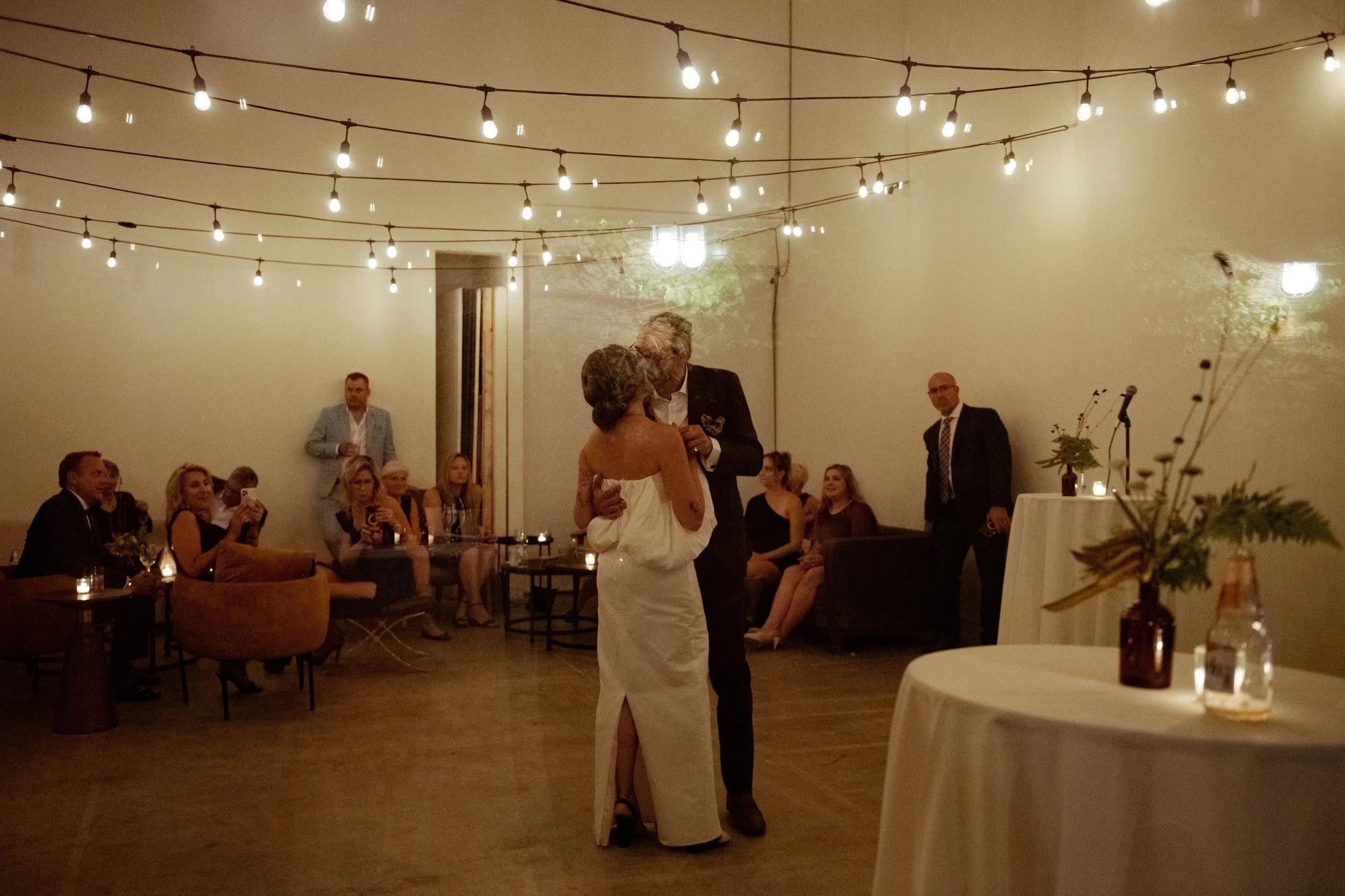 Bride and groom share a dance together underneath string lights and inside a Toronto studio space and event venue rental. Guests sit on nearby sofas and watch the dancing couple. Tables are lit with candles and centrepieces.