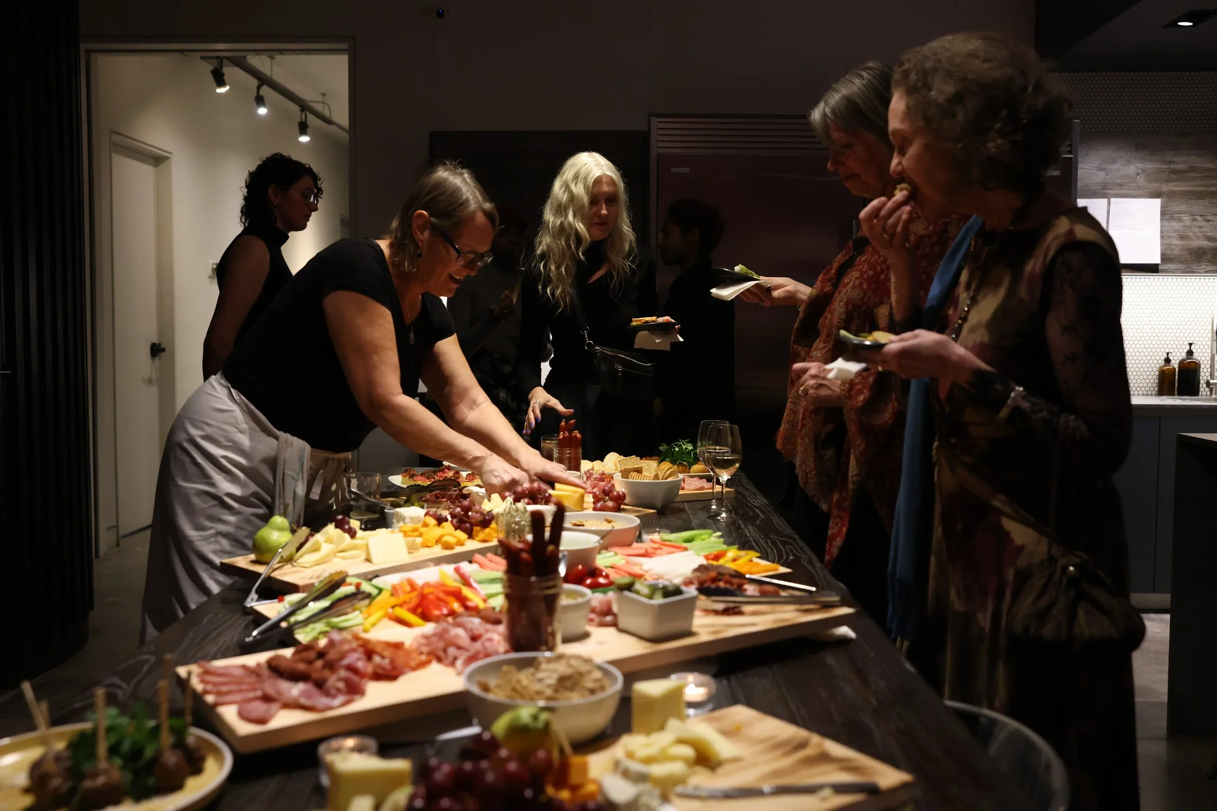 Guests stand around a studio kitchen table and fill their plates with charcuterie.