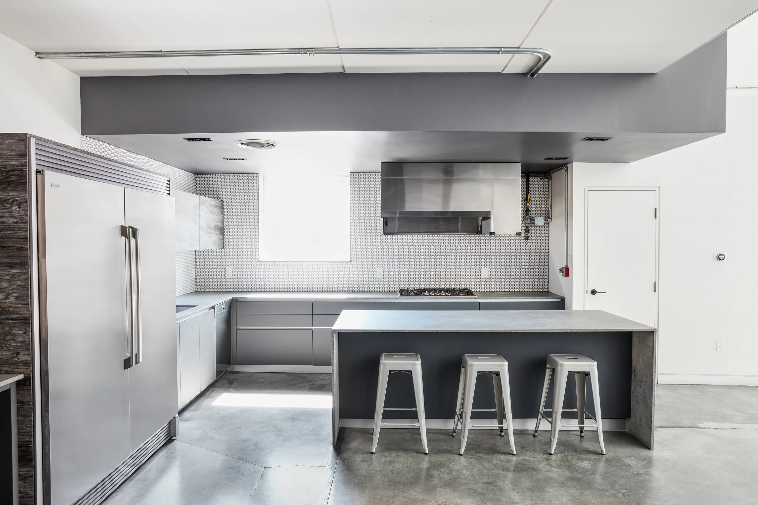 Modern full kitchen island, bar stools, stove range, large refrigerator and freezer, built in dishwasher, cupboards, and countertops.
