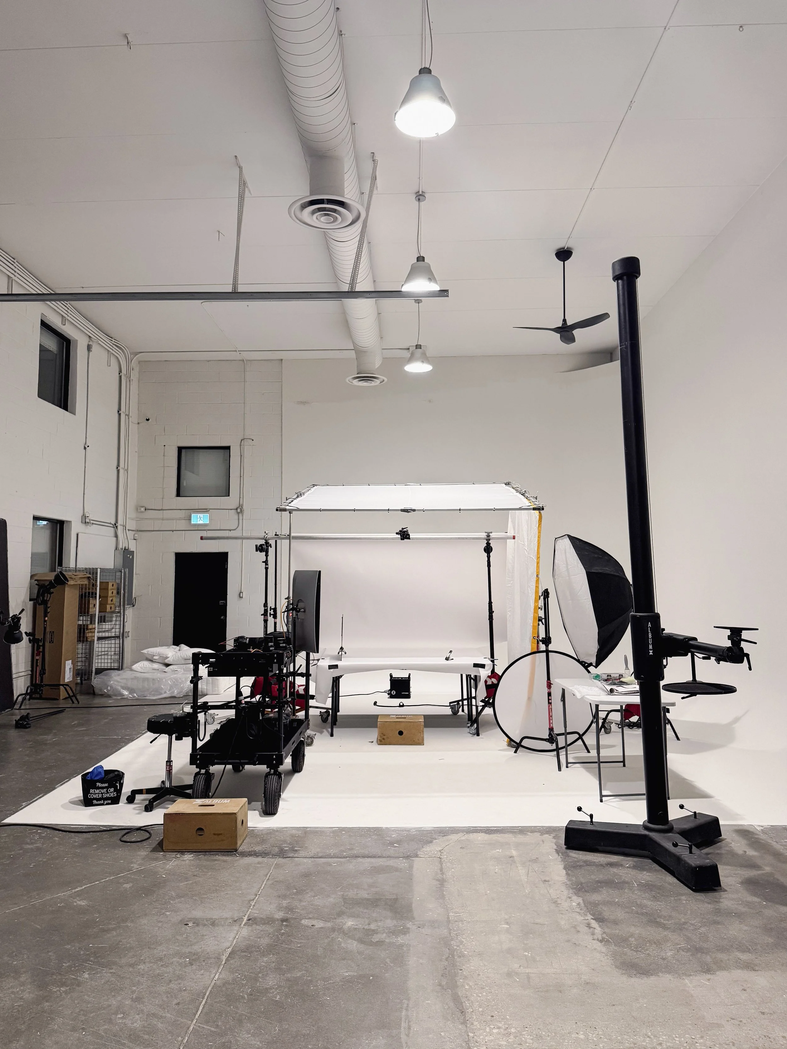 Photoshoot setup on a 28 ft x 19 ft white cyclorama wall. Various photography equipment are on the cyclorama, including apple boxes, Inovativ cart, 12’ Cambo studio stand, reflectors, a monitor, folding table, and sandbags.