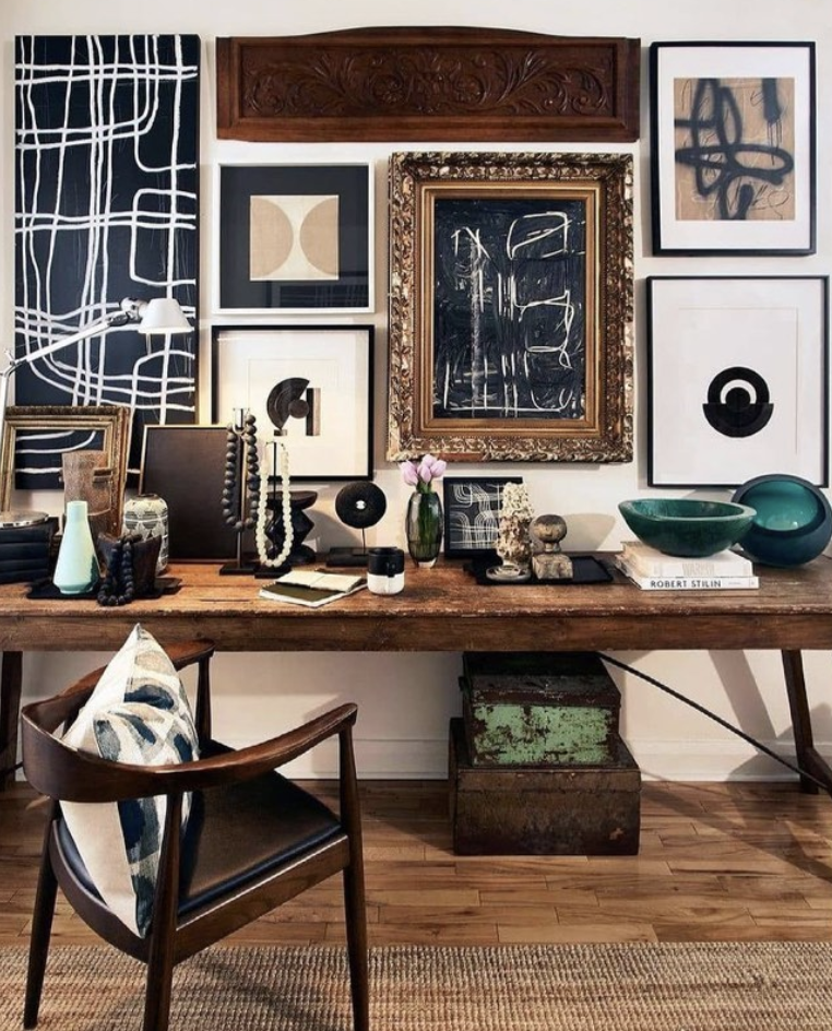 Assortment of paintings on a wall. Vintage office table, wooden office chair, vintage bowls, wood carpet, and books.