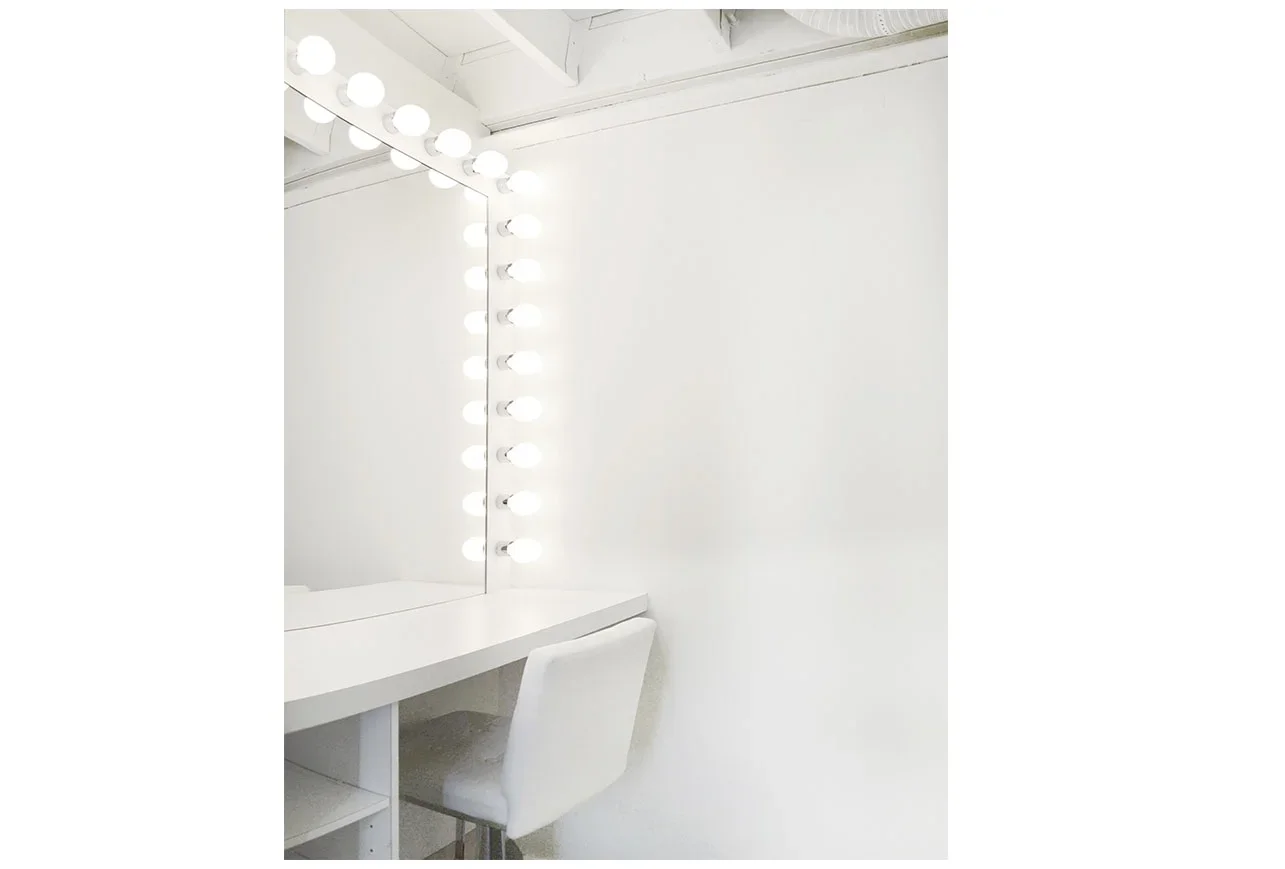 Makeup station with multiple lights framing the vanity mirror. White cushion chair and shelving.