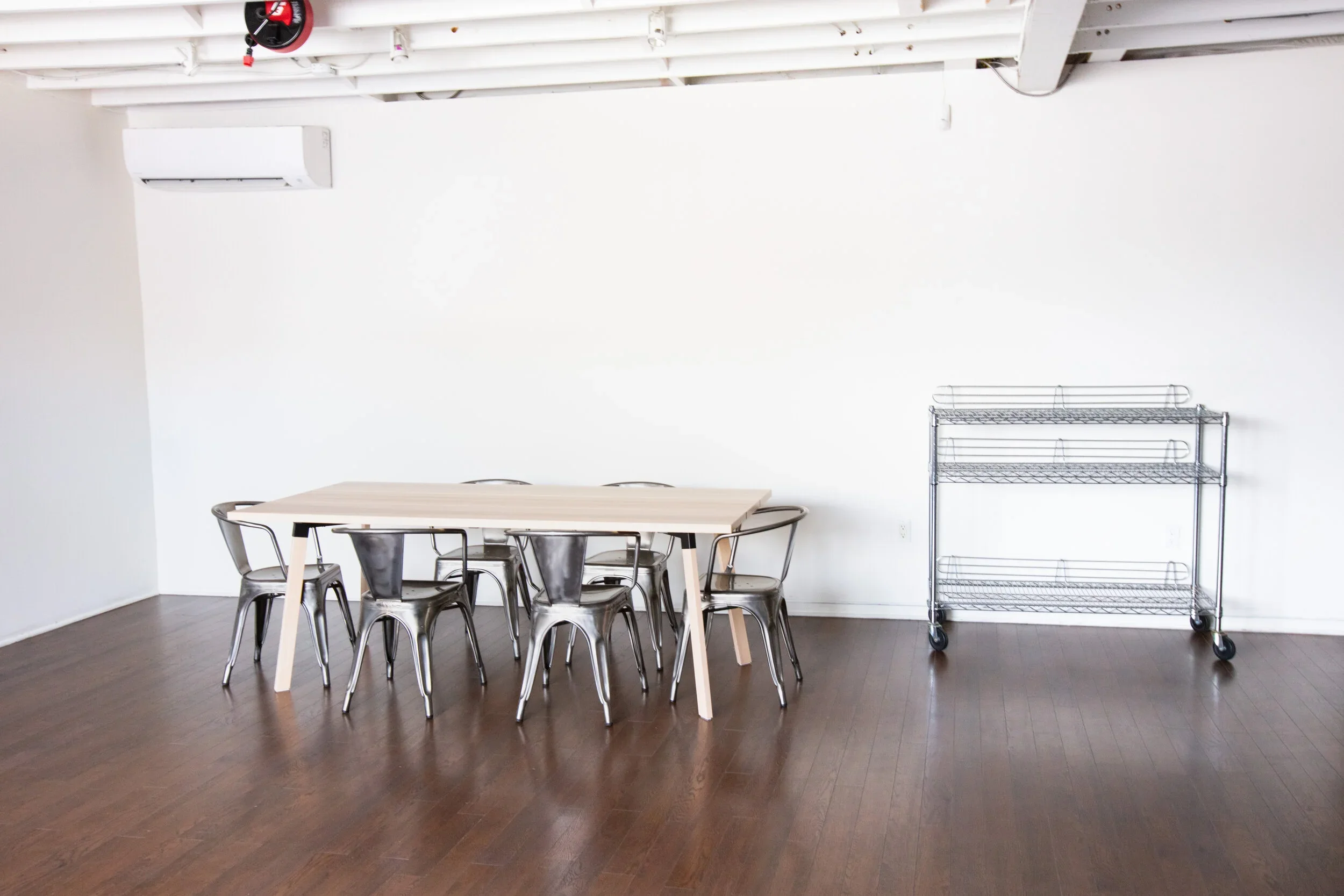 Light wooden table with metal chairs, hardwood flooring, stainless steel cart with wheels, and air conditioner.