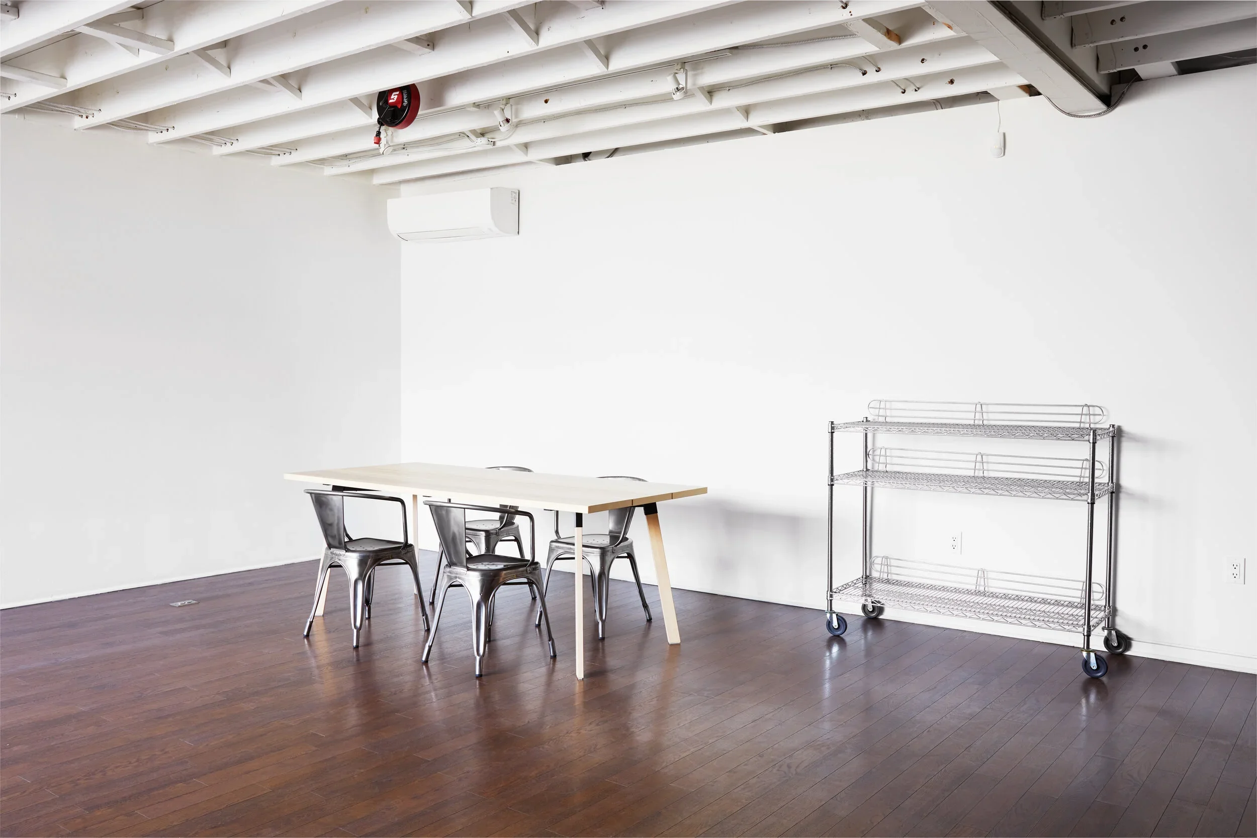 Light wooden table with metal chairs, hardwood flooring, stainless steel cart with wheels, and air conditioner.