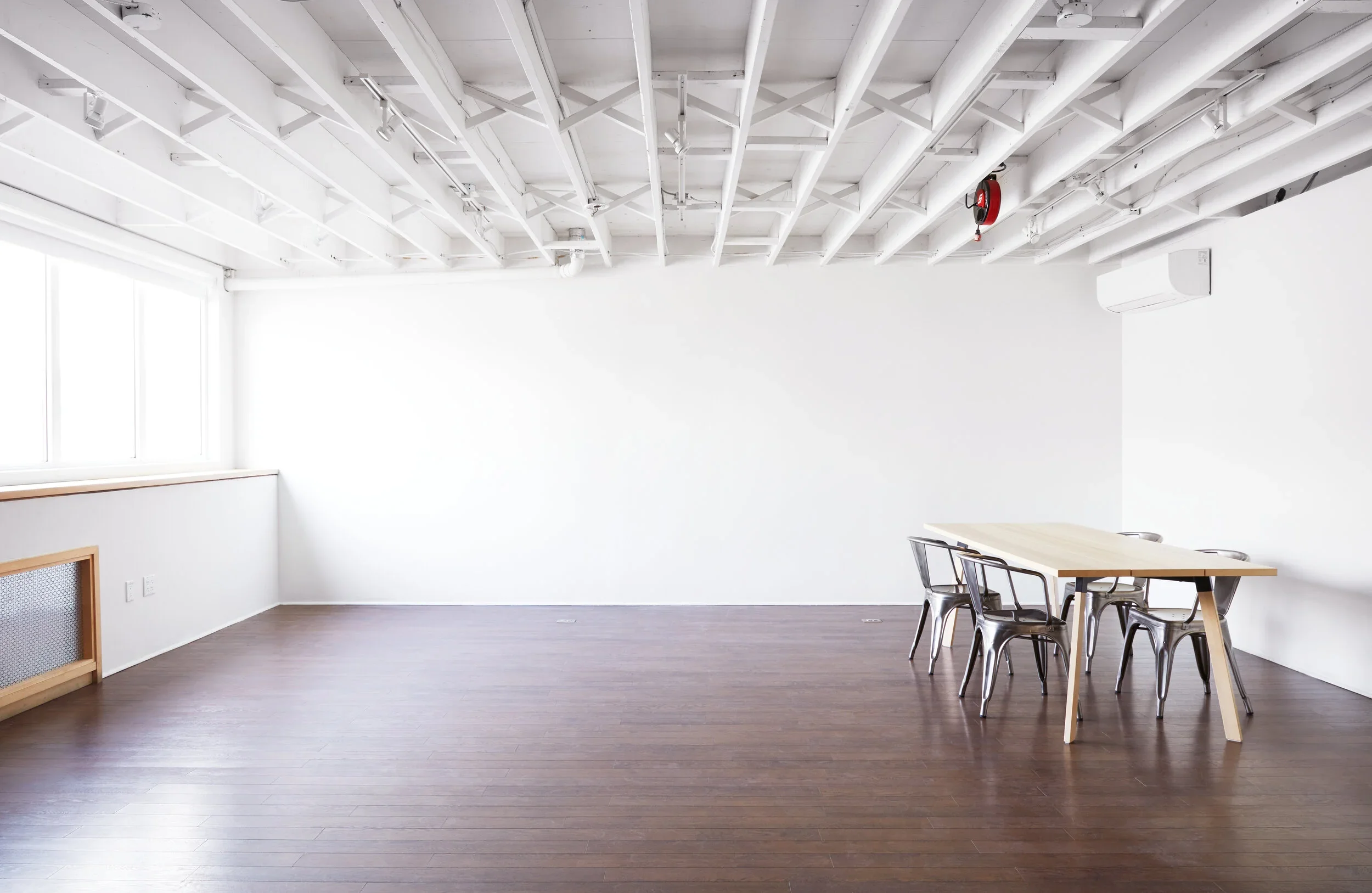 Sunlight coming through east facing windows in a loft style studio. White shooting wall, light wooden table with metal chairs, hardwood flooring, and air conditioner.