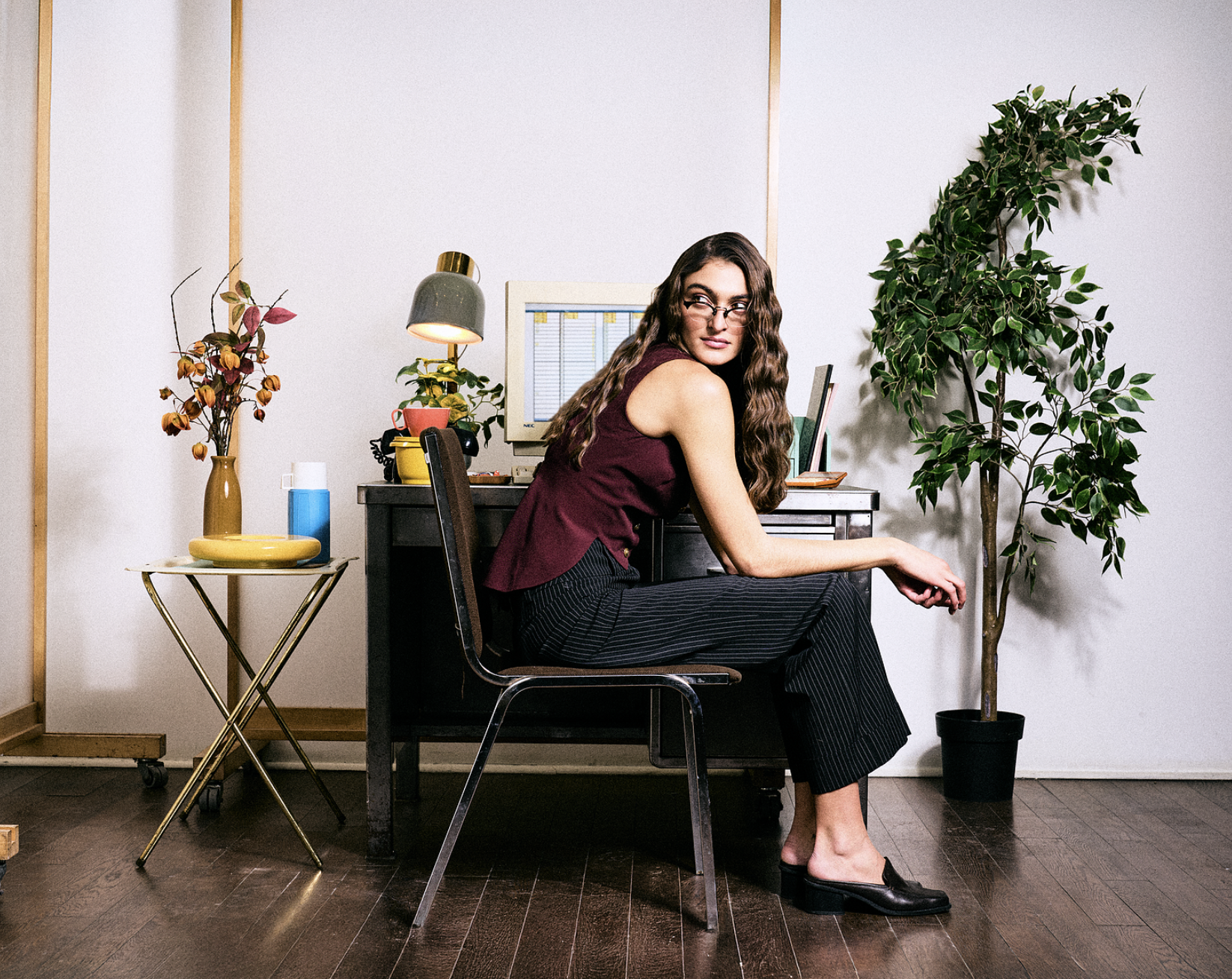 Model sits on a chair and looks over her shoulder. The background shows an office desk with a vintage computer monitor, some plants, and a desk lamp.