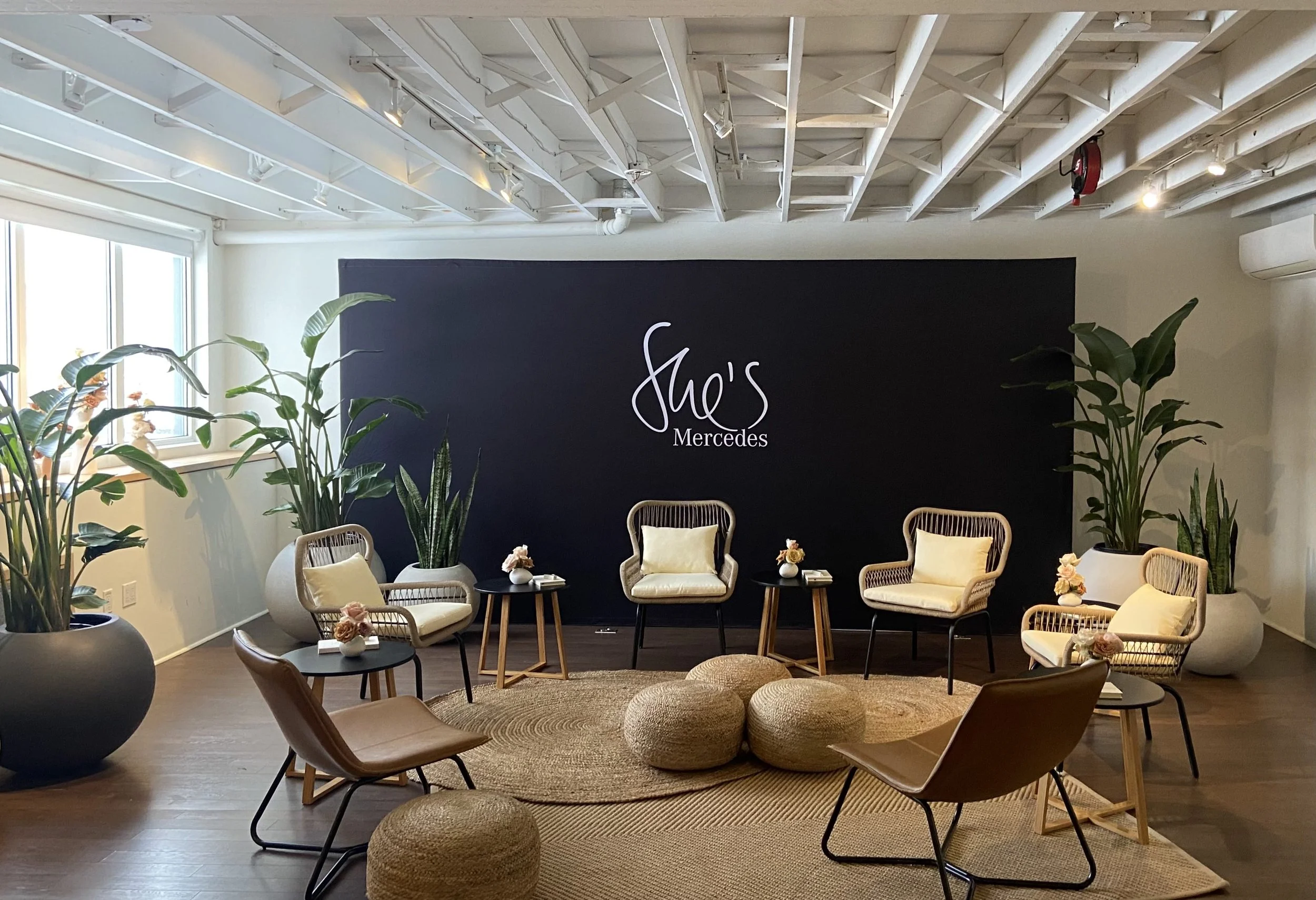 Black backdrop with white text reads 'She's Mercedes' in the background. In the foreground, there are multiple cushioned chairs placed in a circle with brown, stylish rugs. Plants are placed around the studio space.