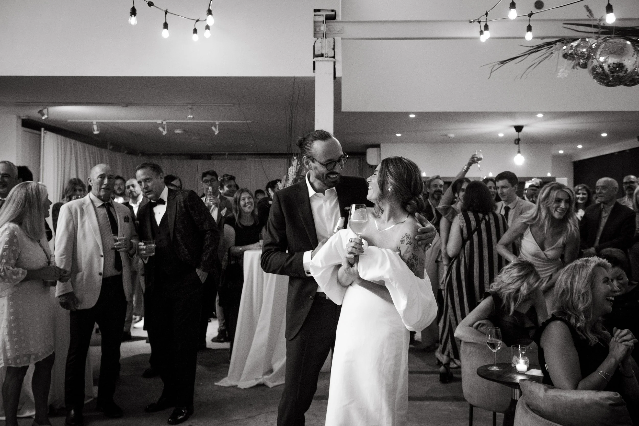 Bride and groom are front and centre and smiling at each other, while a room full of wedding guests interact with each other in the background of a Toronto event venue and studio space.