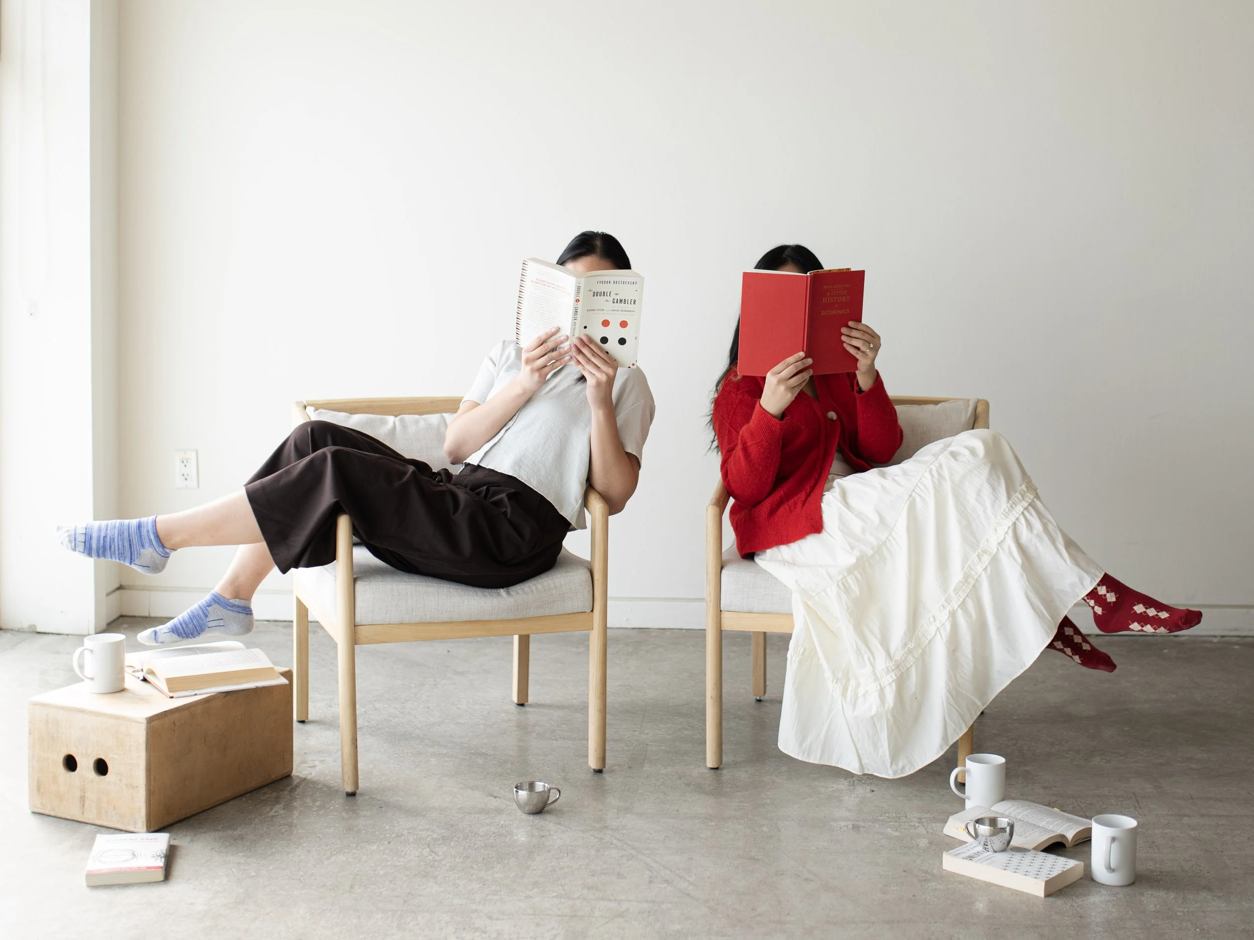 Two models are sitting on chairs next to each other with their faces covered by books. Multiple coffee mugs, books, and apple boxes are scattered on the studio floor.
