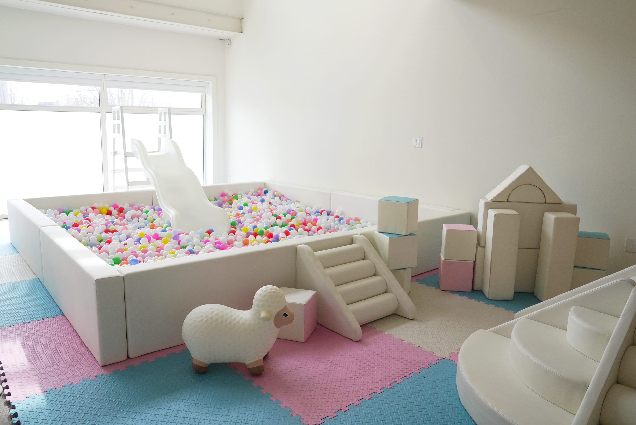 A pink, blue, and white foam mat for children is on the studio floor. There’s a ball pit with a slide, stairs, a life size toy lamb, and foam shapes for playing placed around the ball pit.